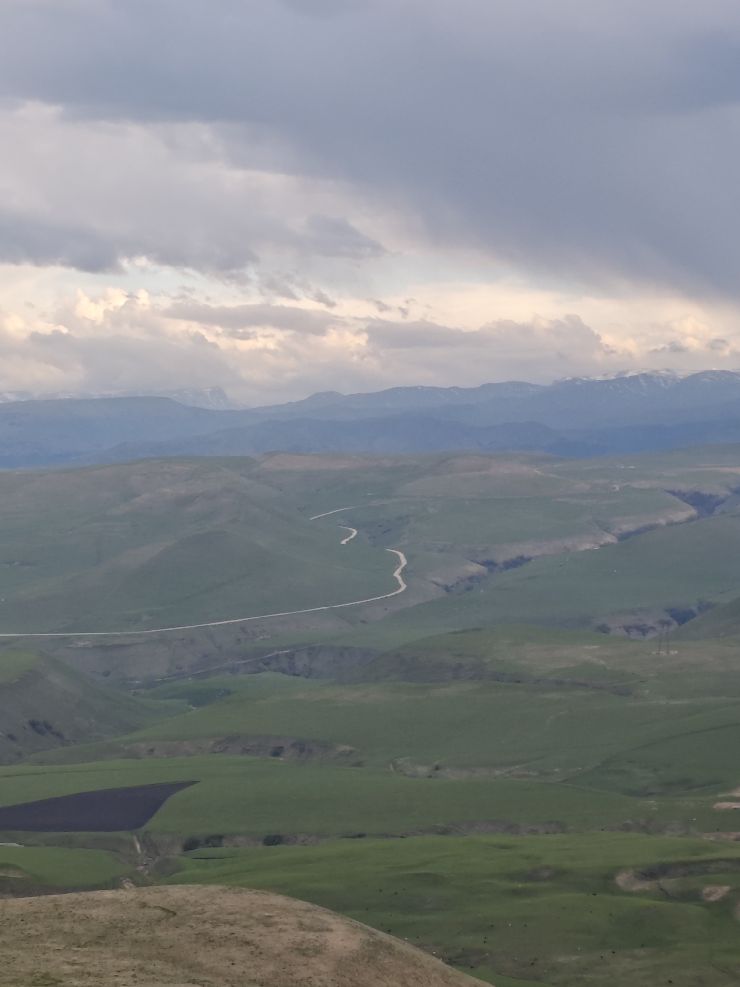 The image depicts a landscape of rolling green hills under a cloudy sky. A road winds through the hills, connecting different areas of the terrain. The distant mountains are visible, with some snow-capped peaks. A dark patch, possibly a field, is noticeable among the green hills. The photograph seems to be taken from an elevated viewpoint. The location is not specified, so the city of Unknown, Unknown is not applicable.