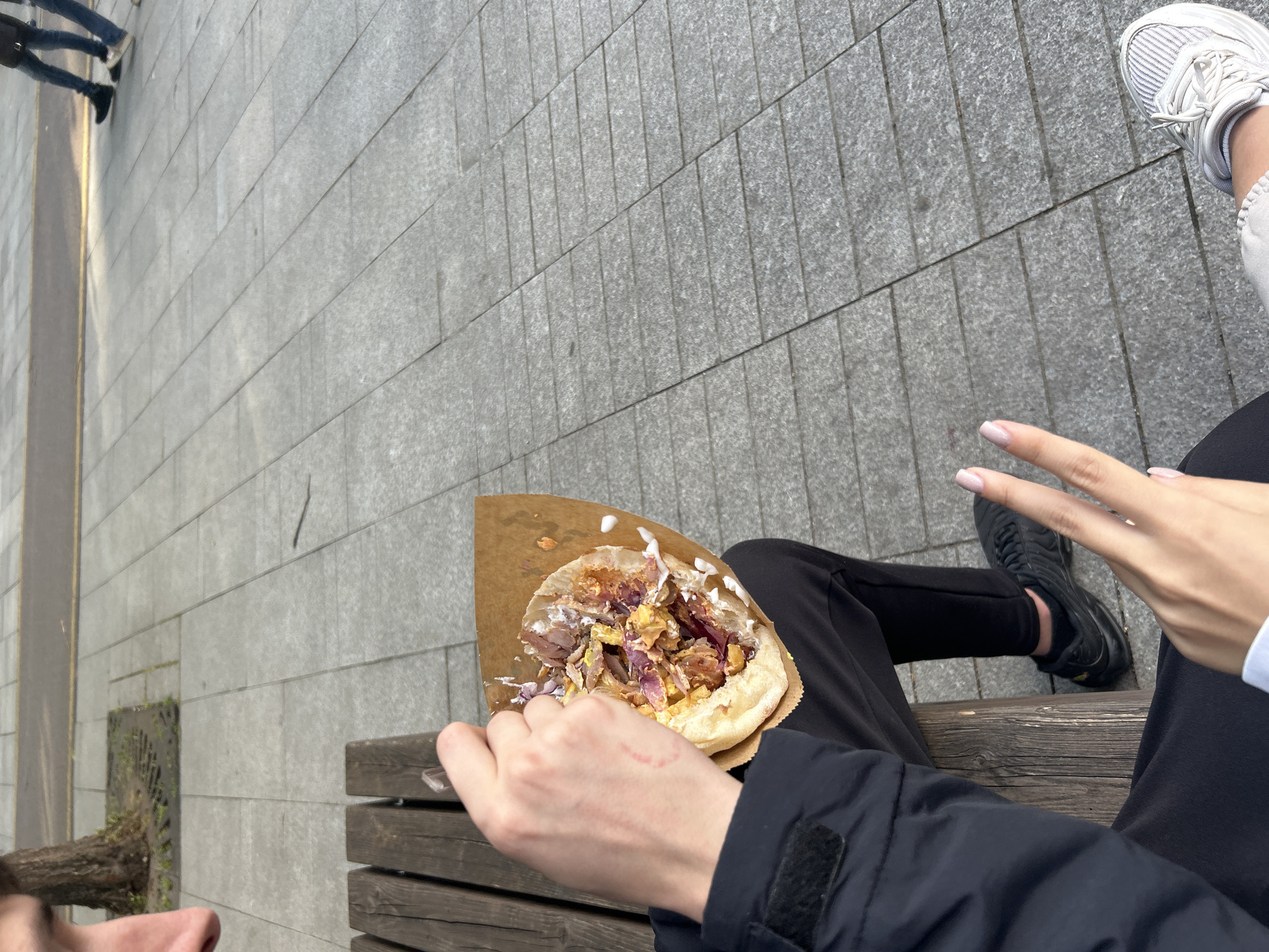 The scene appears to be set outdoors in an urban area, possibly a park or city square, characterized by grey paved ground and a bench.
A person is holding a gyro or similar type of sandwich in one hand, with a brown paper wrapper visible beneath the food. The sandwich is filled with meat, vegetables, and some sort of white sauce.
Another person, wearing black pants and black shoes, is visible partially in the frame. A hand is making a peace sign.
The overall impression is a casual scene, perhaps someone enjoying a snack while sitting on a bench in a city setting.