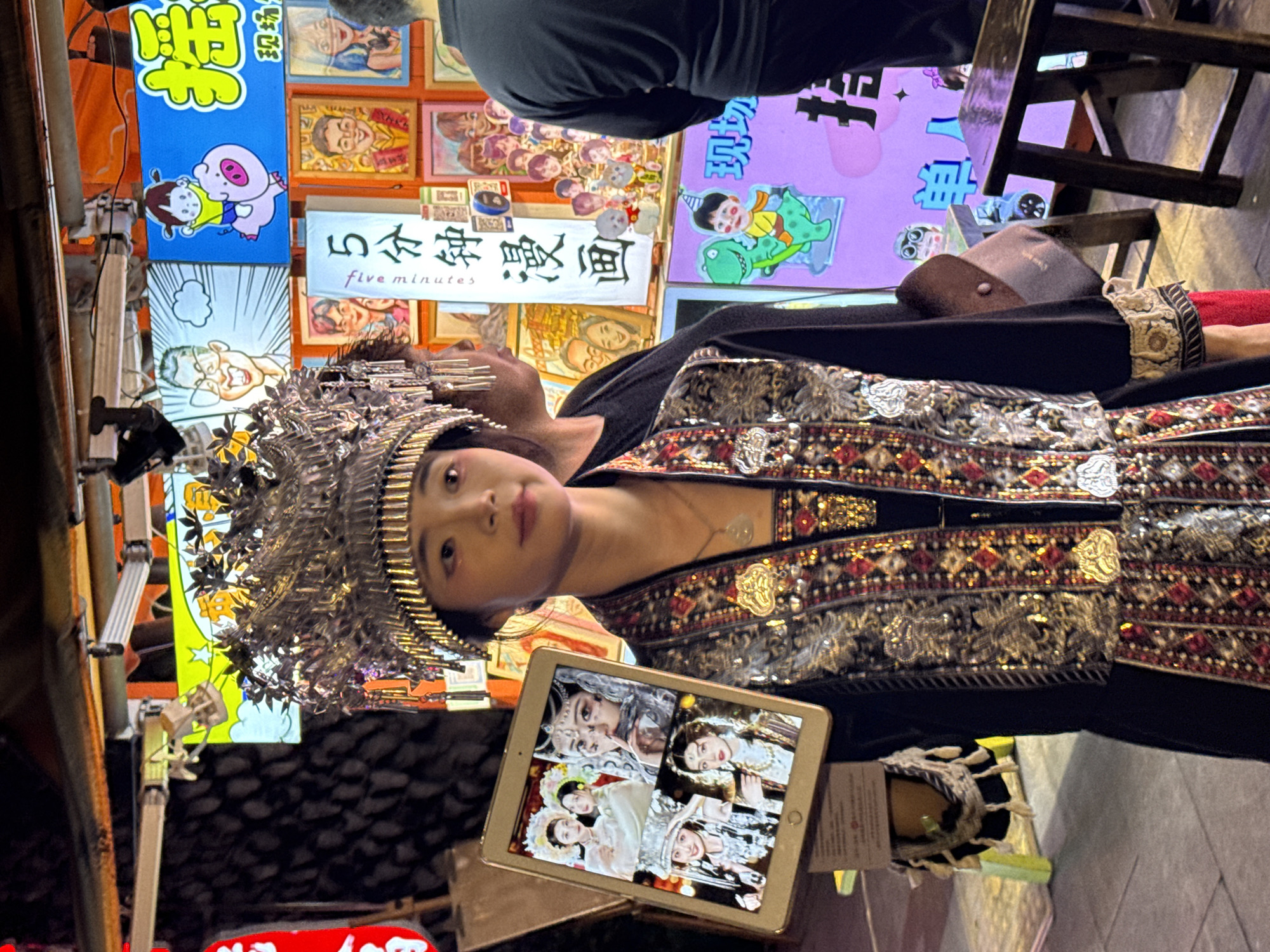 A woman in traditional Miao dress, including an elaborate silver headdress, stands in an outdoor setting in the Yuzhong District of Chongqing, China. She is holding a tablet displaying several images of women in similar Miao attire. Behind her are a series of brightly colored signs with Chinese characters and cartoon figures. A portion of the street and a wooden chair are visible in the background.