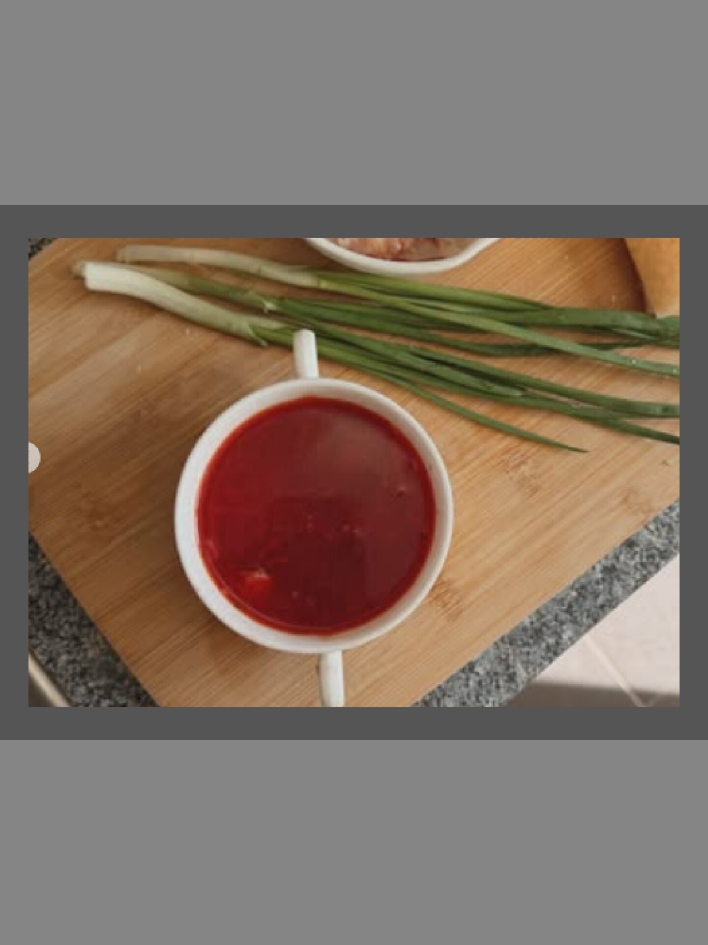 A white mug filled with red soup sits on a wooden cutting board. Next to the mug are several green onions. The background is blurred.