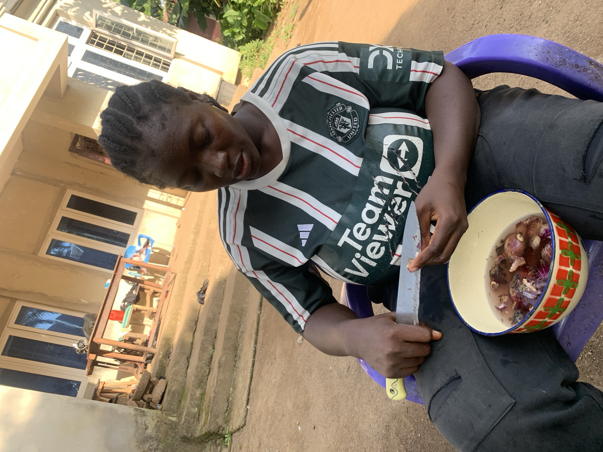 A young woman with braided dark hair sits on a purple plastic chair outdoors in Akwanga, Nigeria, preparing food. She wears a dark green and white striped jersey with "TeamViewer" and the Manchester United logo printed on it, and dark pants. She is holding a knife in her right hand, about to cut into a red onion submerged in a bowl of water filled with other cut vegetables, resting on her lap. The bowl has a colorful geometric pattern around its rim. The background shows a light-colored building with several windows, a wooden table with items on it, and steps leading up to the building. The ground is dirt and sand, and there are some green plants visible. The lighting suggests it is daytime.