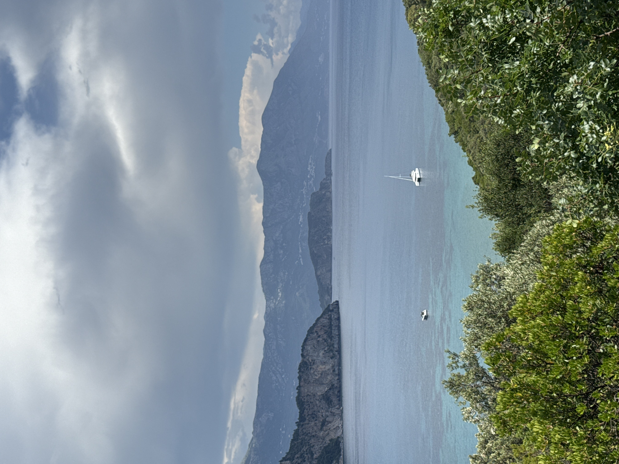 The image depicts a serene coastal landscape with a body of water, mountains, and lush vegetation. In the foreground, vibrant green trees and bushes line a slope leading down to the water's edge. The middle ground features a calm, blue sea with a white catamaran sailing in the distance, as well as another smaller boat. The background is dominated by a range of majestic, hazy blue mountains stretching across the horizon, with a cloudy sky overhead. The scene appears to be a picturesque view of the Adriatic Sea, possibly in or around Dubraljevina, Montenegro, known for its natural beauty and coastal geography. There are no people visible in the image.