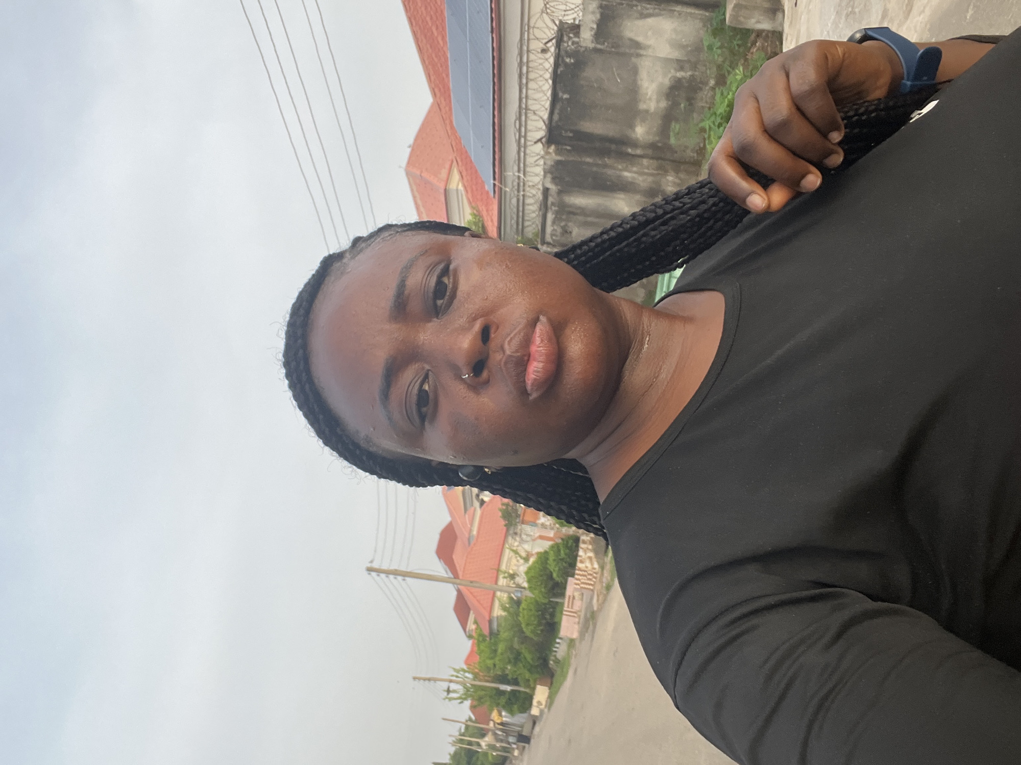 A selfie shows a Black woman with braided hair, wearing a black shirt and a blue smartwatch, holding the end of her braid in her right hand. She has a nose ring and appears to be outdoors. The background includes power lines, buildings with orange roofs, a concrete wall topped with barbed wire, and trees. The location appears to be a residential area in a city, potentially in Abuja, Nigeria.