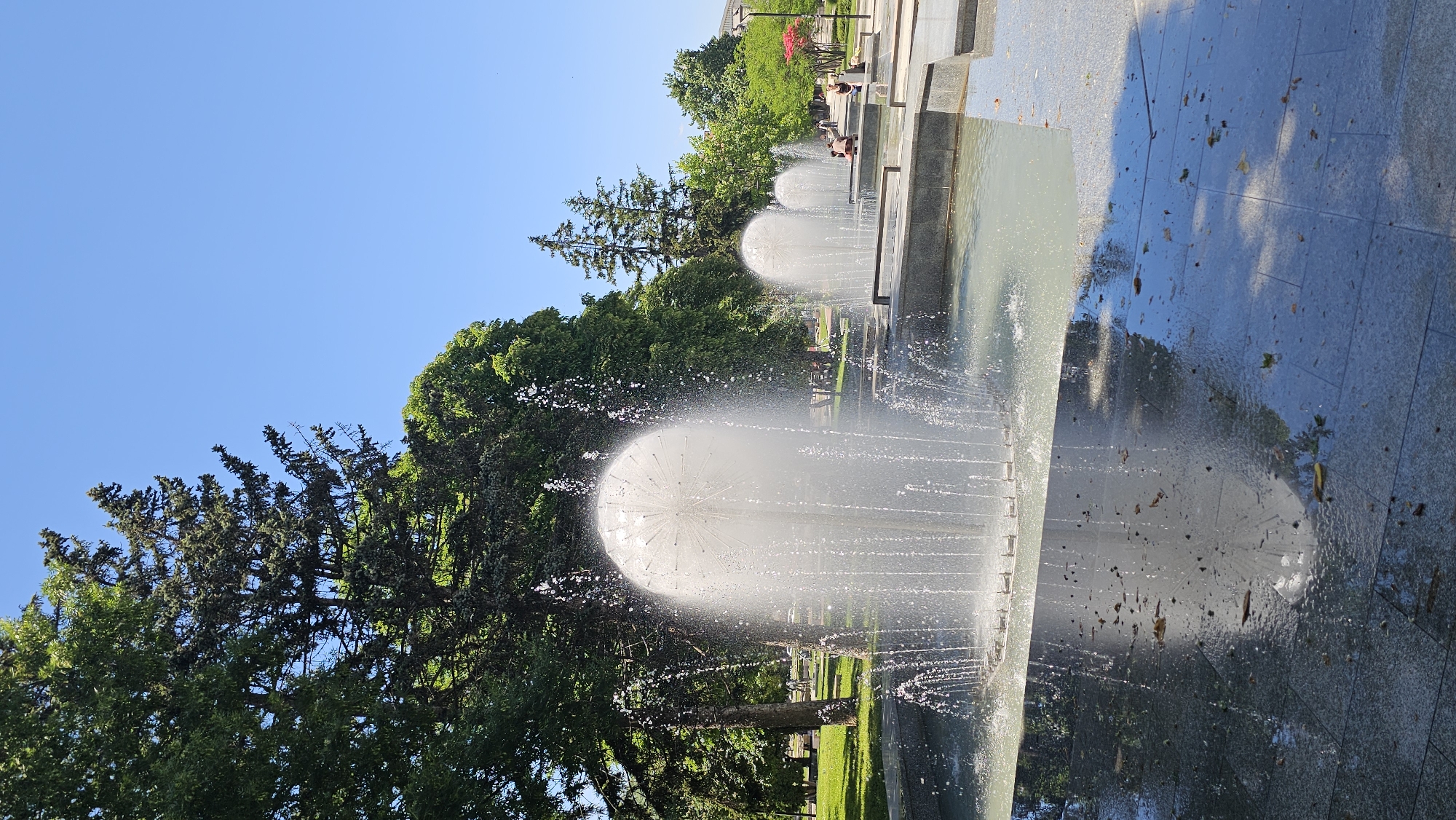 A low-angle, outdoor shot depicts a series of fountains against a backdrop of trees and a clear blue sky. The fountains are configured with water jets forming spherical shapes. The fountains are set within concrete structures with a tiled surface that appears wet and reflective. There are some individuals in the background sitting near the edge of the fountains or the structures containing them. The trees, predominantly green, provide a natural backdrop. The sky above is a clear, cloudless blue.