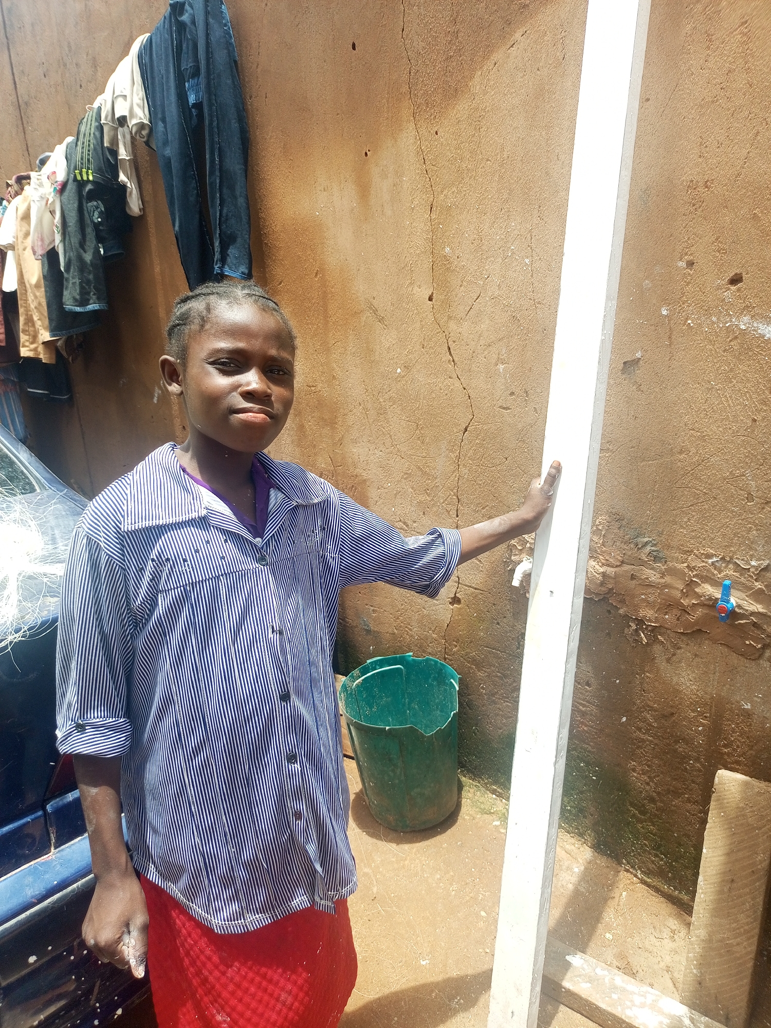 In Tempure, Nigeria, a young person stands outside next to a textured, earth-toned wall. They are wearing a blue and white striped button-down shirt and a red skirt. A white vertical beam is positioned to their right, and they are holding onto it with their right hand. Behind them, clothes hang on a line, and a blue vehicle is partially visible. In the foreground, a green bucket sits on the ground, and a blue faucet is mounted on the wall to the right of the beam. The ground appears to be dirt or sand.
