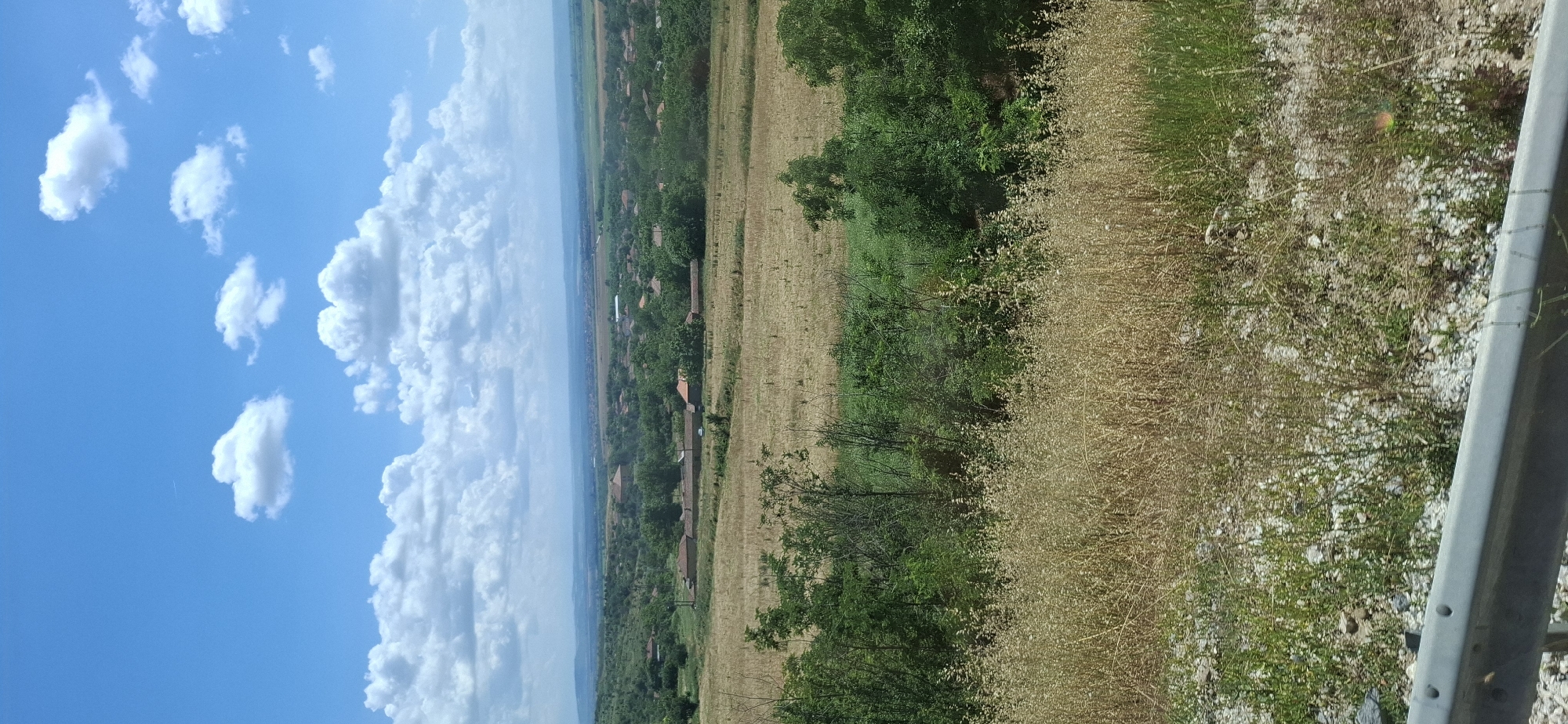 The image depicts a landscape view of a rural area, likely a village, under a partly cloudy blue sky. In the foreground, a metal guardrail is visible with gravel and sparse vegetation. The middle ground features fields of dry, golden grass and patches of green trees. Beyond that, a cluster of houses with red-tiled roofs, typical of Bulgarian architecture, are scattered across rolling green hills. The scene suggests a sunny day with a vast expanse of nature and human habitation. The metadata indicates the location is Kozar Belene, Bulgaria. There are no people or animals visible in the frame, nor any discernible actions or interactions. The overall environment is tranquil and bucolic.