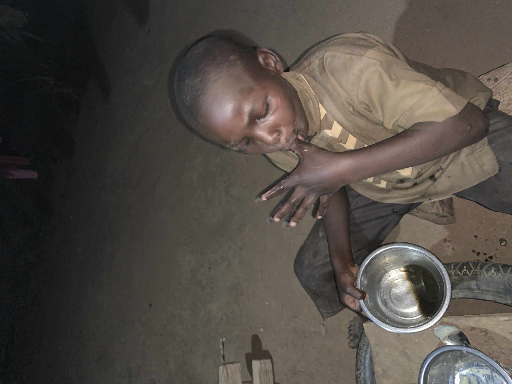 A boy is shown from an overhead angle, licking his fingers as he holds a metal bowl containing a dark liquid. The environment appears to be a dimly lit dirt floor area, possibly indoors. Additional objects in the scene include another metal bowl and a partially visible tire. The location provided is Agban, Nigeria.