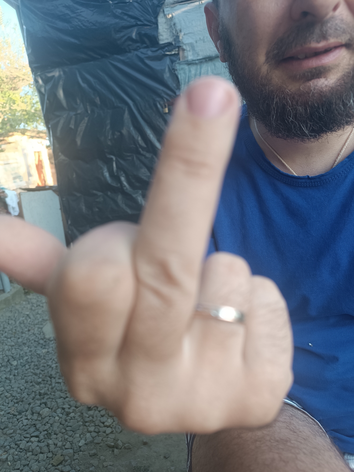 In Vypasne, Ukraine, a person with a beard and wearing a blue shirt is holding up their middle finger to the camera. The background consists of a rough, possibly unfinished structure covered in black plastic sheeting, with some vegetation and gravel visible. A simple silver ring is worn on the person's middle finger. The image appears to be a close-up shot, with the hand and part of the face being the primary focus.