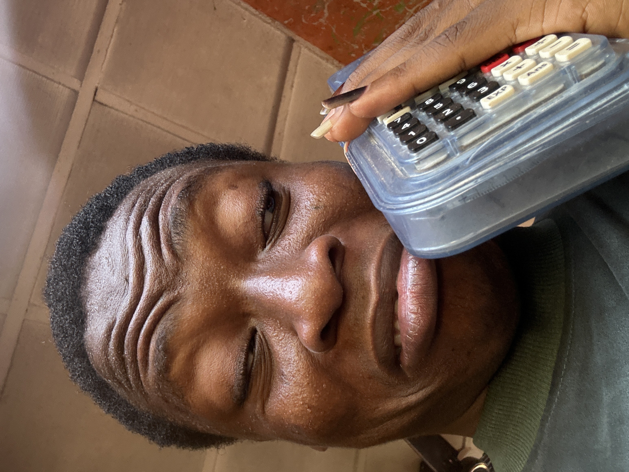 A close-up, rotated portrait features a person with dark skin and short, curly dark hair, positioned with their face oriented to the right of the frame. The perspective is upward-looking. The person's forehead displays prominent creases, and their eyes are partially closed or squinting, suggesting an expression of concentration or slight strain.
A hand, also with dark skin, is visible in the upper right, holding a transparent plastic device. The hand has long, light-colored fingernails with dark polish on the tips. The device resembles a small calculator or similar electronic gadget, with numerous keys visible through its clear casing. Key labels include numerical digits (e.g., 1, 4, 7, 0, 2, 5, 8, 3, 6, 9) and function keys like "EXP," "Ans," "X," and "+," some of which are colored red or white/cream.
The person is wearing a green ribbed collar, indicative of a shirt or sweater. The background is a light brown or tan textured surface, possibly an indoor wall or ceiling, given the unusual camera angle. The activity appears to be the person intently using or interacting with the handheld electronic device.