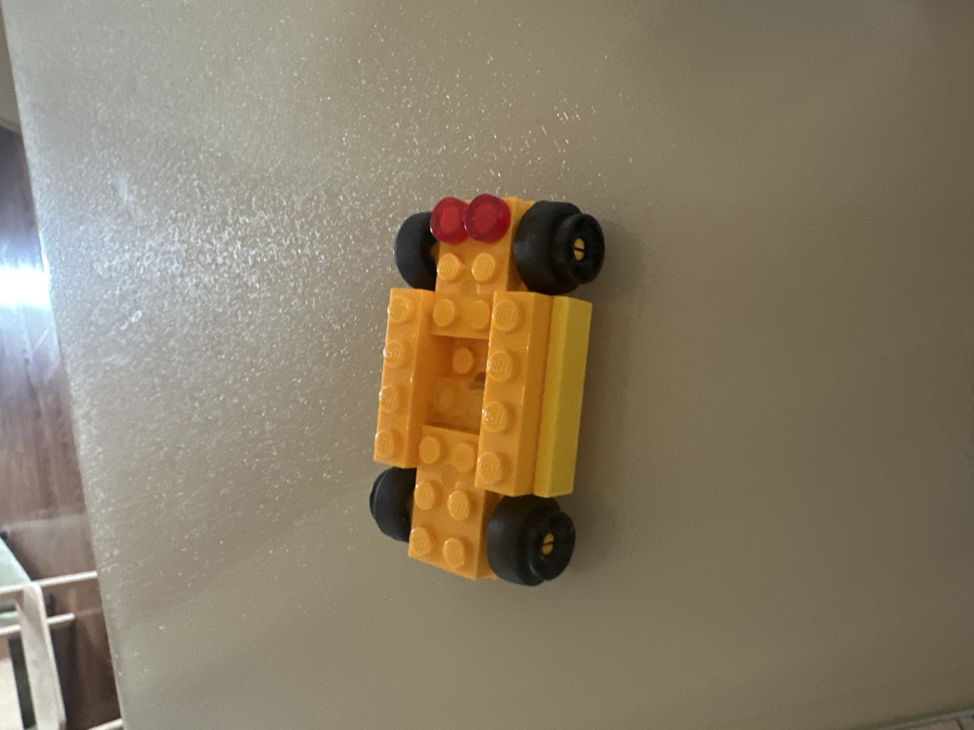 A small, orange and yellow Lego car is resting on a light-colored, textured surface. The car has four black wheels and two red taillights. The background shows a blurred wooden floor and what appears to be shelving. The image is taken from a top-down perspective. There is no visible indication of the city of Kiparisnoe, Ukraine.
