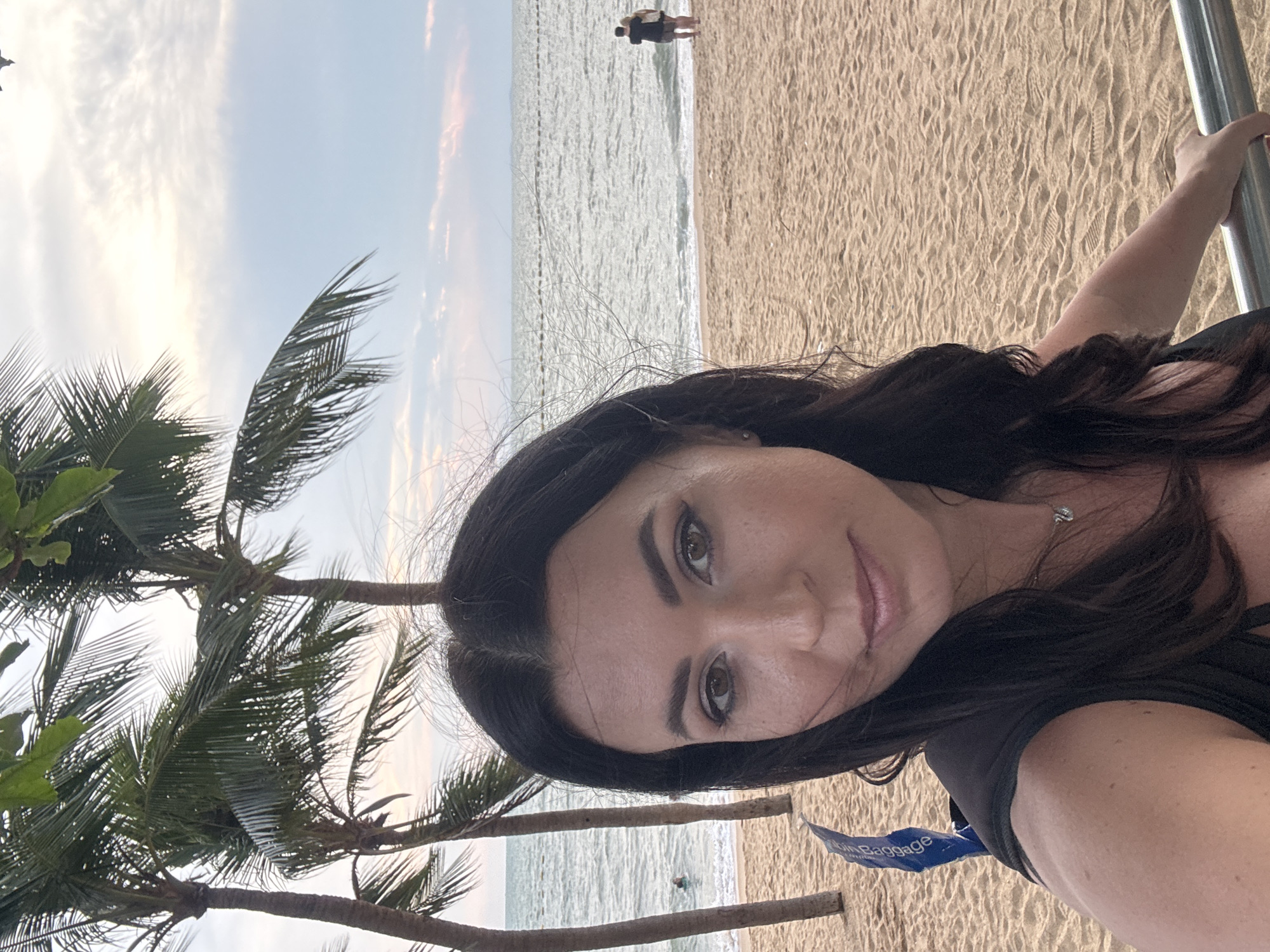 A selfie captures a woman posing outdoors at a tropical beach location, likely Pattaya City, Thailand. The woman, with long dark hair and brown eyes, is in the foreground, looking directly at the camera with a calm expression. She is wearing a dark-colored top and a delicate silver necklace. Her right arm is visible, holding onto a silver metal pole or railing.
Behind her, the scene transitions to a sandy beach adorned with footprints, leading to the ocean with gentle waves lapping at the shore. Further in the distance, a solitary figure can be seen standing on the beach or wading in the shallow water. To the left side of the frame, several tall palm trees with green fronds are prominent, adding to the tropical atmosphere. The sky above is bright but soft, with light clouds, suggesting a late afternoon or early evening setting, possibly during the golden hour. A small blue tag with white text "Cabin Baggage" is visible near the woman's right arm.