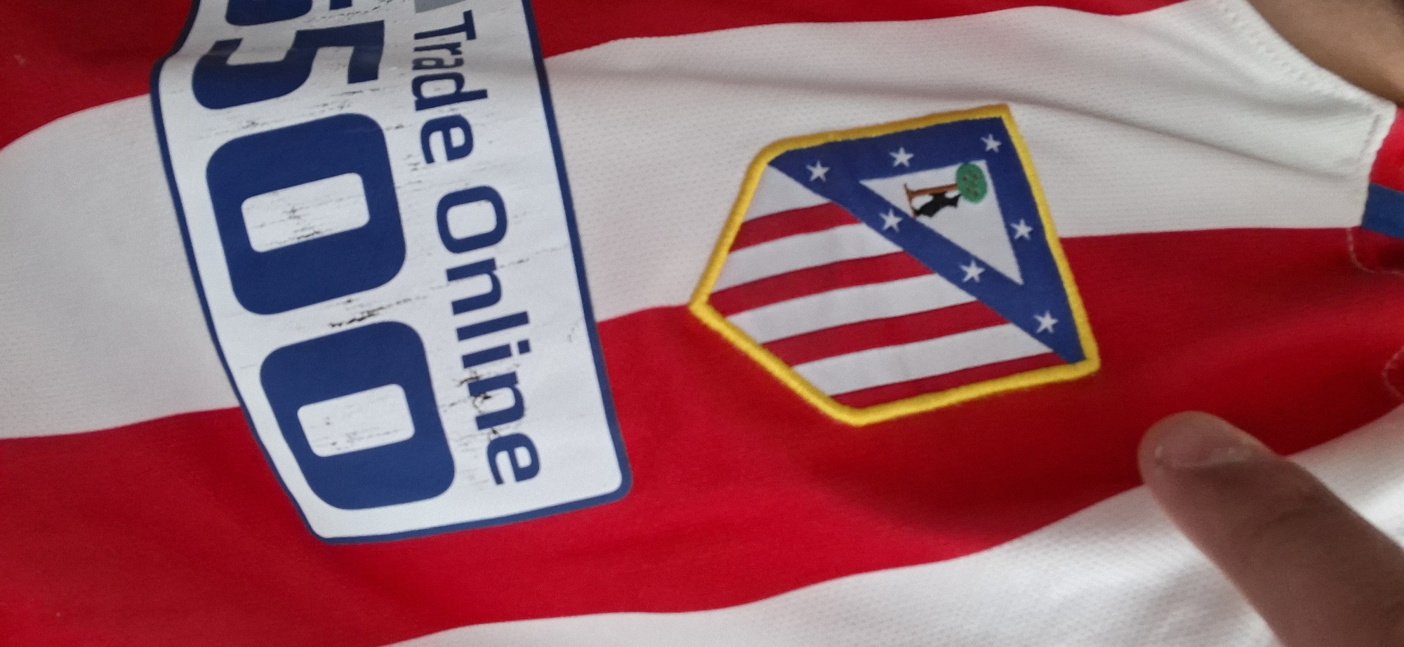 A close-up shot captures a red and white striped sports jersey, likely a football (soccer) shirt, with a visible sponsor logo and club crest. The jersey features prominent vertical red and white stripes.
On the left side of the image, running vertically, is a white rectangular sponsor patch with blue text. The text "500 Trade Online" is clearly visible, although the "500" part is more prominent, and the lettering appears slightly distressed or worn, suggesting a printed design rather than embroidery.
To the right of the sponsor logo, a hexagonal club crest is embroidered onto the jersey, outlined in yellow. The crest is divided into two main sections. The top-left triangular portion is blue with several white stars and depicts a brown bear standing on its hind legs, reaching for a green tree with a red trunk. The bottom-right section of the crest features red and white vertical stripes.
A light-skinned finger is visible in the bottom-right corner of the frame, pointing towards a red stripe on the jersey, below the club crest. The fabric texture of the jersey is discernible, characteristic of athletic wear. The image is taken at an angle, providing a dynamic view of the details on the shirt. There are no other specific location cues within the image beyond the jersey itself.