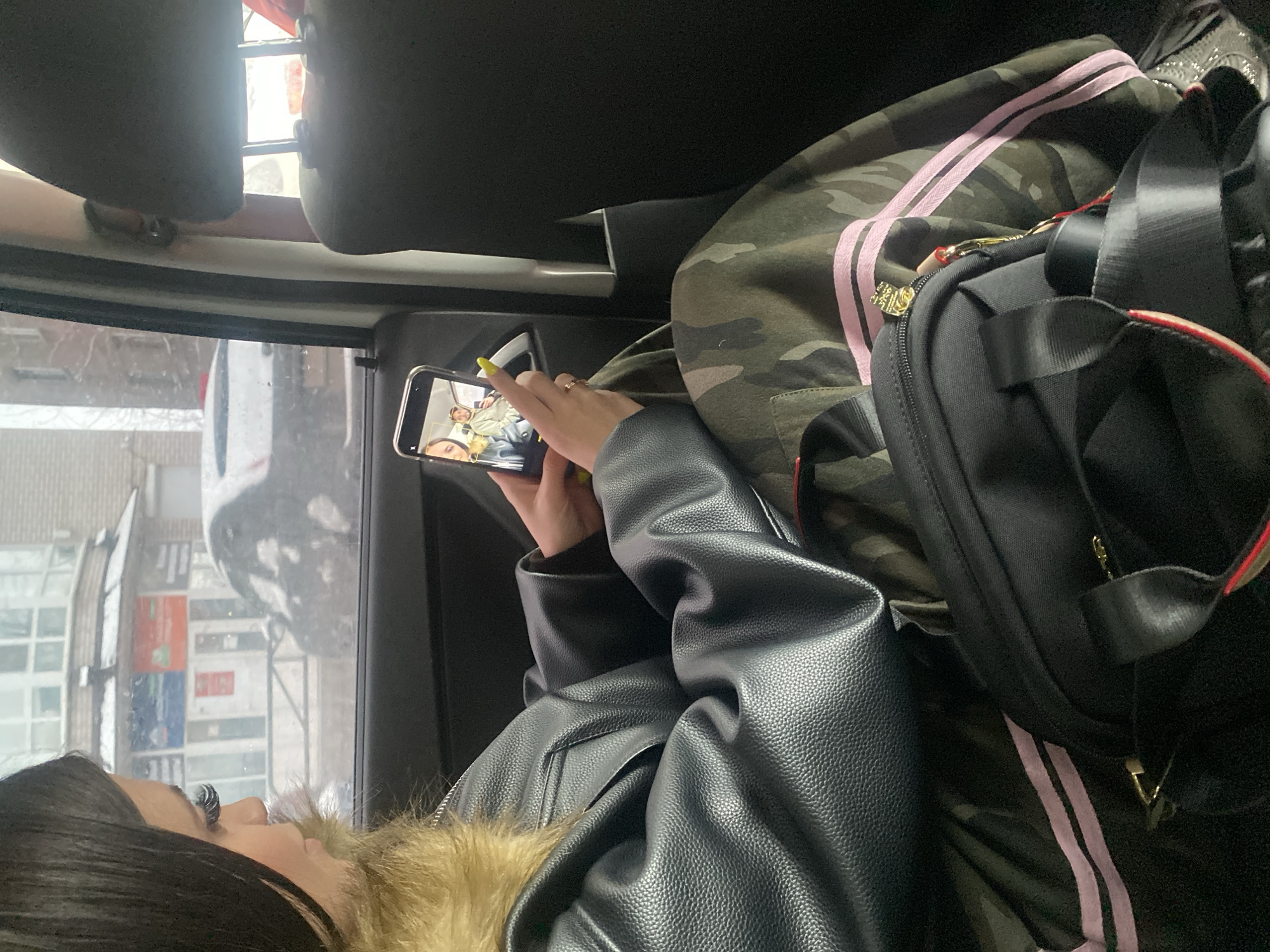 A woman sits in a car, viewing a phone screen. She wears a black leather jacket with fur trim, and has long black hair. On the screen of her phone, two people, presumably in the same car, are visible. Next to her is a camouflage-patterned tracksuit with pink stripes and a black bag. Outside the car window, snow is visible on the ground and on the car's side mirror. Buildings are seen through the window. Based on the buildings, including a distinctive blue and red facade, the location is Chaikovsky, Russia.