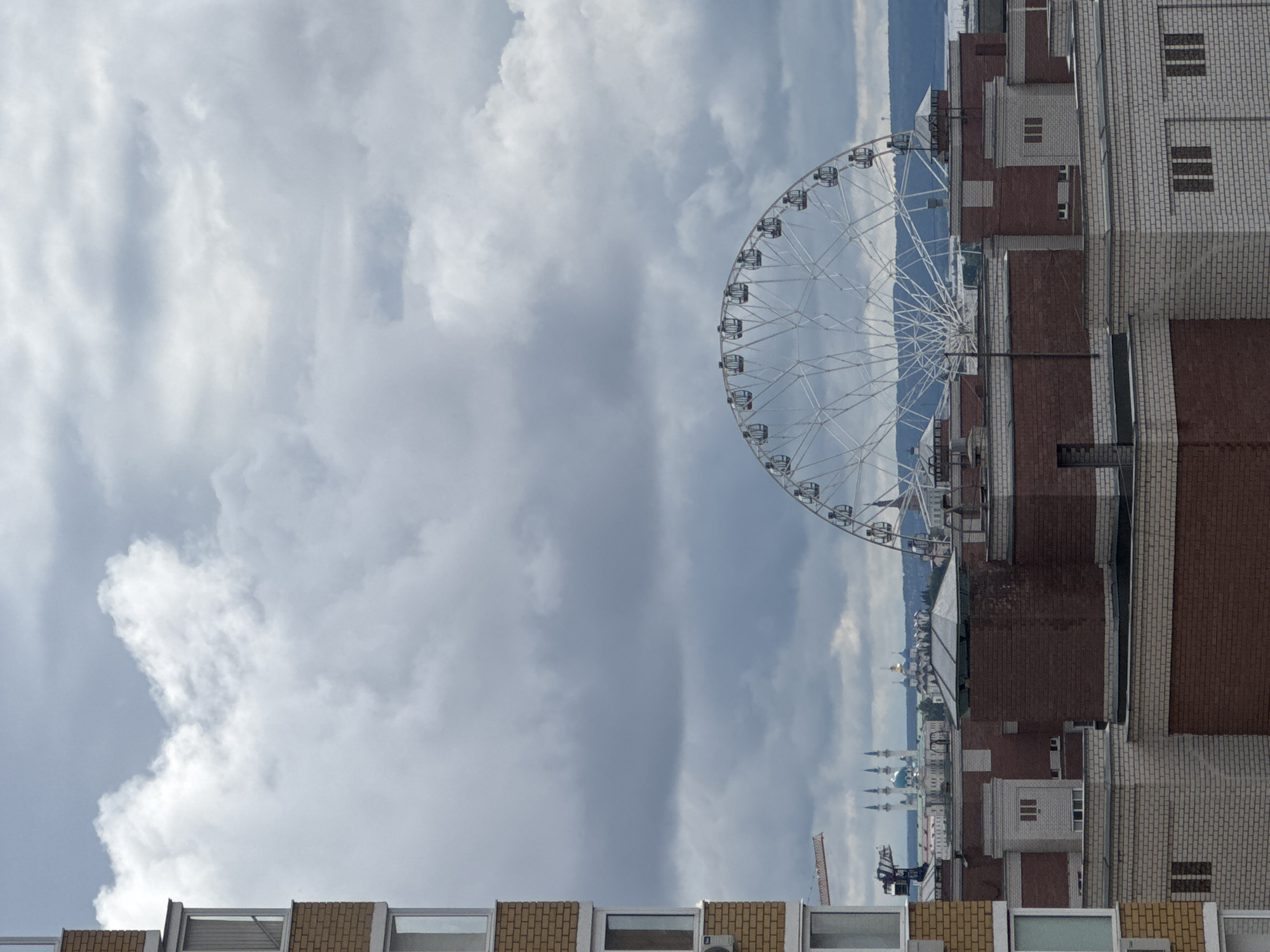 The image shows a Ferris wheel positioned beside a brick building under a cloudy sky. The Ferris wheel is white and appears modern. The brick building is primarily brown and white with visible windows. A portion of another building is visible in the lower part of the image. In the background, there's a cityscape with structures including minarets and domes. The location is the city of Kazan, Russia.