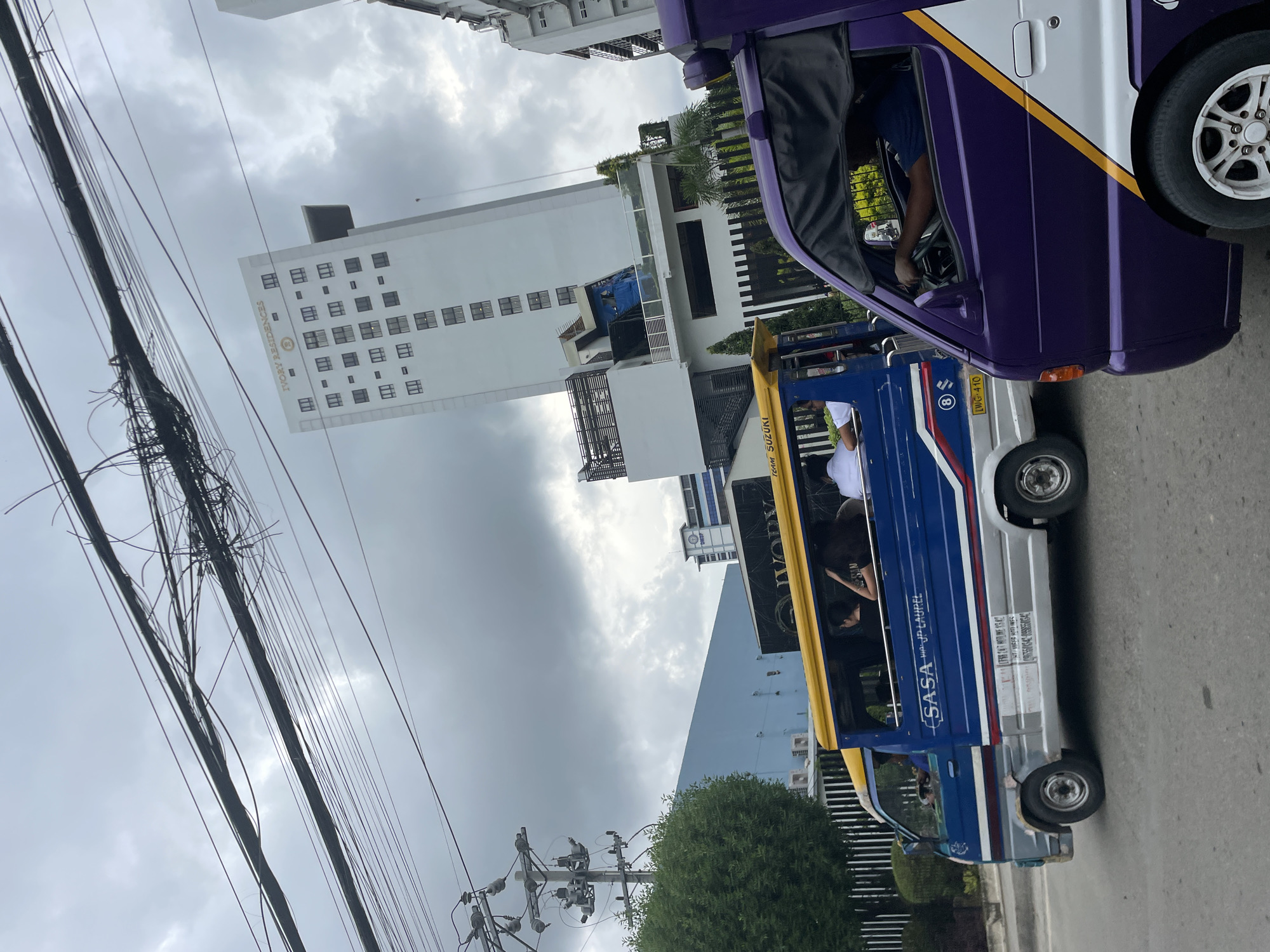 This is an eye-level shot of a street scene in Davao City, Philippines, under an overcast sky. The foreground features the side view of a purple tricycle. Behind it, a blue and silver jeepney, a common mode of public transportation in the Philippines, is traveling. The jeepney is decorated with the text "SASSA" and "JP. LAUREL" along its side, indicating its route. The interior of the jeepney shows several passengers, including women with their hair down and a man in a white shirt.
In the background, a tall, white building with many windows stands prominently, identified by the text "IVORY RESIDENCES" at its top. The building appears to be a residential complex. To the left of the building, there is a tall utility pole with numerous tangled wires, a common sight in urban areas of the Philippines. Further left, a lush green tree provides a contrast to the urban elements. The street itself is paved, and other vehicles and buildings are partially visible.