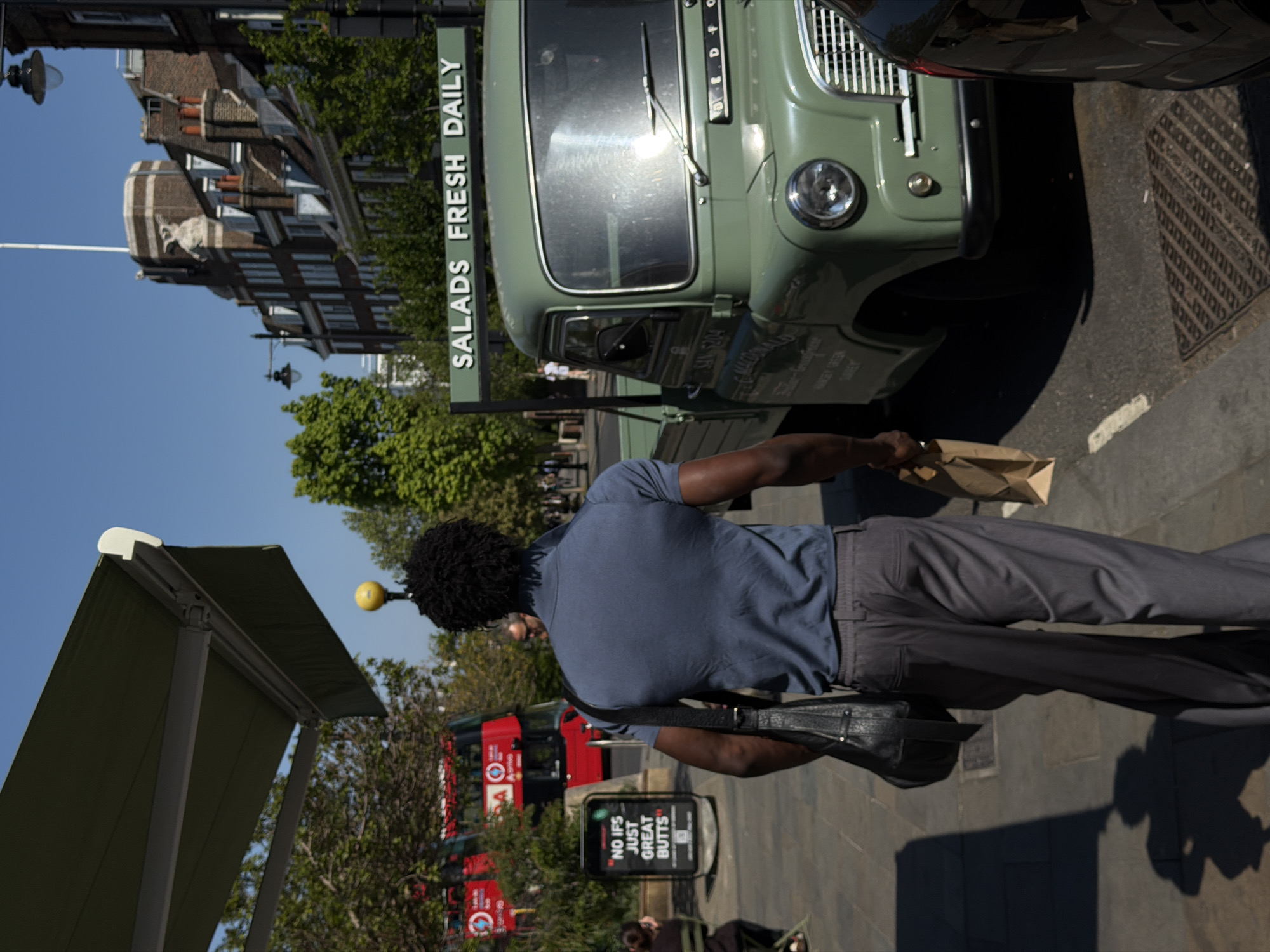 The image captures a scene in London, United Kingdom, showing a man in a blue t-shirt and gray pants walking on a sidewalk. He is carrying a brown paper bag and a black shoulder bag. To his right, a vintage green "Salads Fresh Daily" food truck is parked on the side of the road. Behind him, red double-decker buses can be seen in the background. A sign that reads "No ifs, just great butts" is also visible. In the upper left corner, there are brick buildings. The sky is clear and blue, suggesting it is daytime.