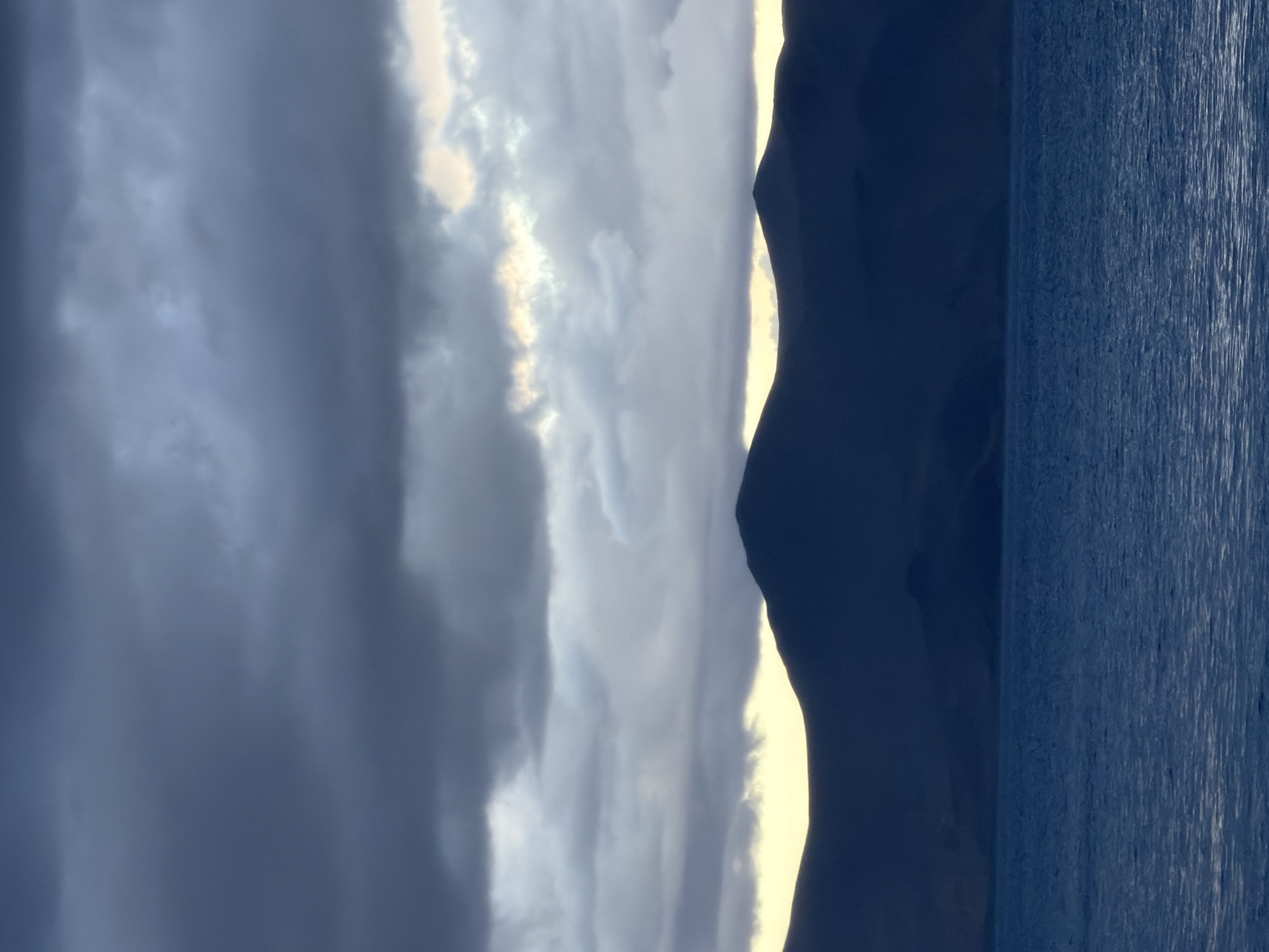 A silhouette of a mountain range is visible across a body of water, with a dusky sky filled with dark clouds overhead. The mountains, rendered in deep blue, are stark against a faint yellow glow that peeks through the clouds near the horizon. The water in the foreground is a rich, dark blue, with ripples suggesting a gentle breeze. The overall scene evokes a sense of calm and the grandeur of nature, typical of the high-altitude landscapes around Karzok, India, specifically the Pangong Tso lake.