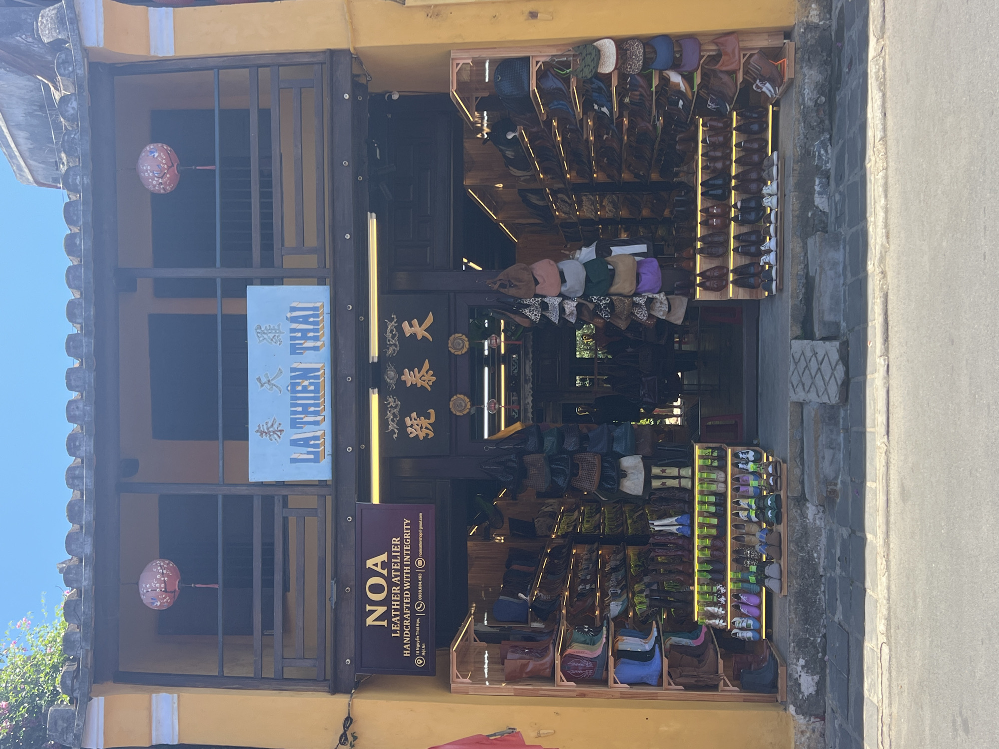 The image shows the exterior of a shop in Hội An, Vietnam. The shop appears to be a leather atelier named "NOA LEATHER ATELIER" as indicated by a sign above the entrance. The shop is located on a street with a paved sidewalk and a road in front. The building is painted yellow and has a traditional architectural style with a wooden balcony. Two red lanterns are hanging from the balcony. Inside the shop, there are numerous leather products, including boots and bags, displayed on shelves. The street is clean and the sky is clear and blue, suggesting a sunny day. No people are visible in the image.