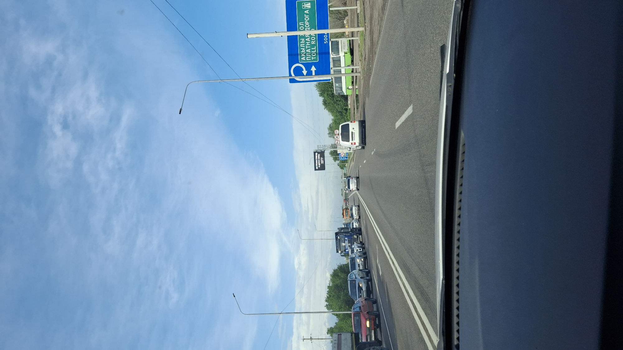 The image shows a highway scene in Панфилово, Kazakhstan. Traffic is heavy, with cars and trucks lined up on the road. A large blue sign indicating "Toll Road" is visible, along with overhead power lines and street lights. The sky is blue with scattered clouds.