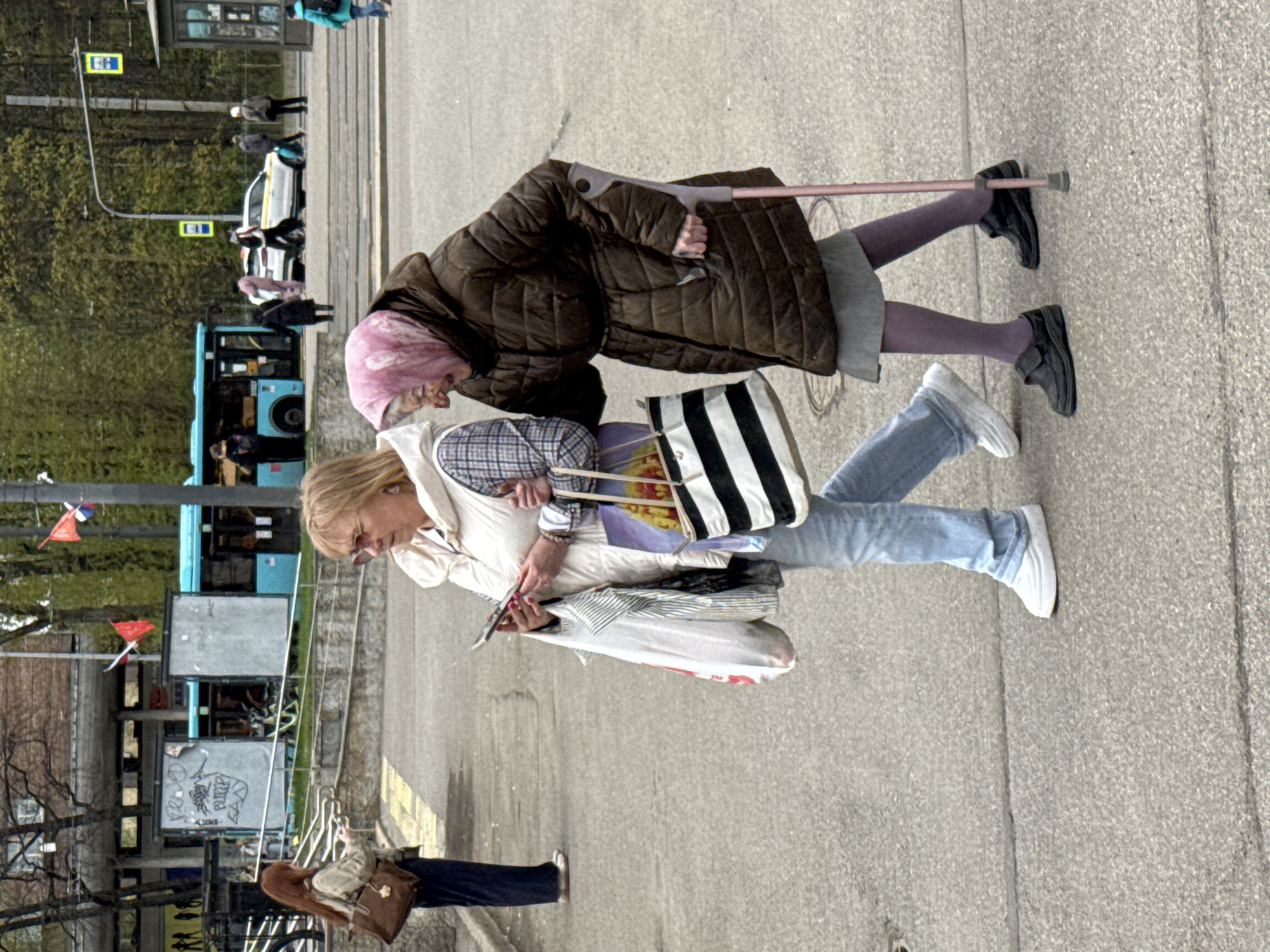 Two women are walking together on a paved street. One woman is older, wearing a brown puffy coat, purple leggings, black shoes, and a pink headscarf. She is using a crutch for support. The other woman is wearing a white jacket, light blue jeans, and white sneakers. She is carrying several shopping bags and holding a smartphone. A blue bus is visible in the background, and there is street signage and trees in the distance. The scene is likely in Saint Petersburg, Russia, given the red, white, and blue flags visible. A third person is walking away in the lower left corner.