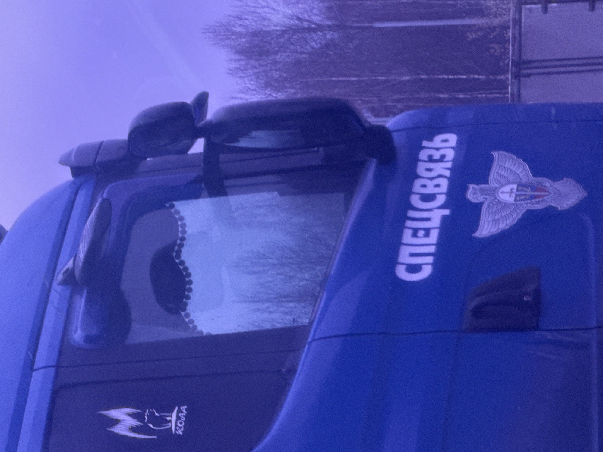 The image displays the passenger side of a blue truck cabin, viewed from an outdoor perspective. A side window with a decorative scalloped curtain is visible, along with multiple side mirrors positioned near the window and along the roofline. Prominently featured on the truck's blue panel is the vertical white Cyrillic text "СПЕЦСВЯЗЬ" (SPETSSVYAZ), translating to "Special Communications" or "Special Mail Service." Adjacent to this text is a detailed emblem, likely representing an official service, possibly depicting a stylized winged figure or a double-headed eagle with a shield. Lower on the cabin, another emblem is visible, featuring a stylized animal, possibly a wolf or horse, accompanied by the Cyrillic text "KONA." The background, seen through the window and above the truck, shows bare trees against an overcast sky, suggesting a winter or early spring setting. The entire image is bathed in a distinct purple-blue color cast. The Cyrillic text is consistent with the provided location context of Klenovskoe, Russia. No specific activity or event is captured; the truck appears to be stationary.