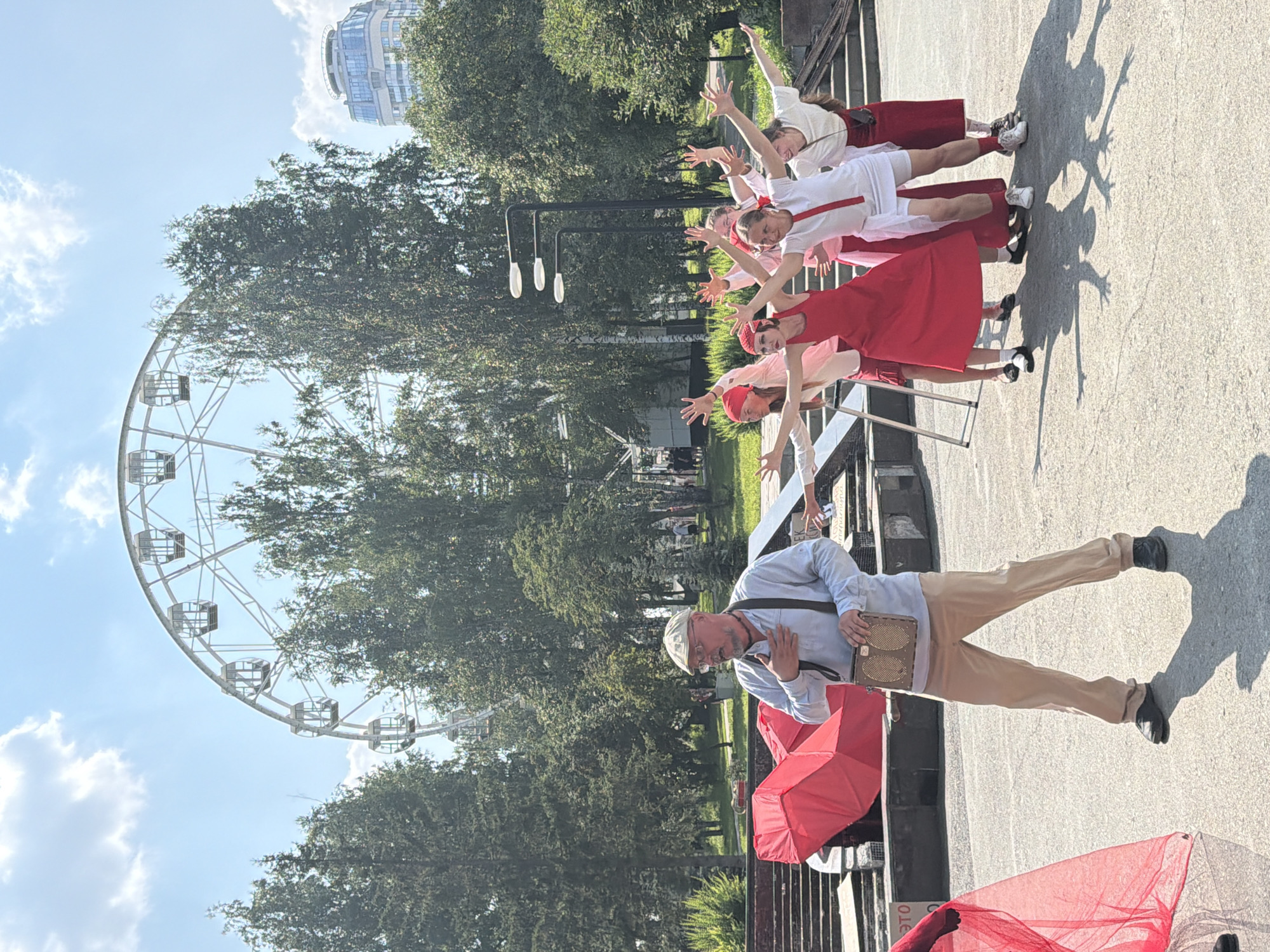 In Yekaterinburg, Russia, under a bright, partly cloudy sky, a group of performers is engaged in an outdoor theatrical or dance presentation near a Ferris wheel. The Ferris wheel, a prominent feature of the landscape, is visible in the background, along with trees and a modern building.
In the foreground, a man in a light blue shirt, tan trousers, and a cap stands with his hand on his chest, looking to his left. He wears glasses and has a satchel across his shoulder. To his right, a group of women, dressed in various shades of red and white attire, are performing with outstretched arms, their faces turned upwards and outwards, suggesting a synchronized movement or pose. One woman in the center wears a bright red dress, while others are in white tops and red skirts or trousers. The ground is paved with a textured surface, and the shadows of the performers are cast distinctly on the ground. A large, red, umbrella-like structure is partially visible to the left of the man, and a red flowing fabric is also present on the far left edge of the frame. The overall scene suggests a public performance in an urban park or recreational area.
