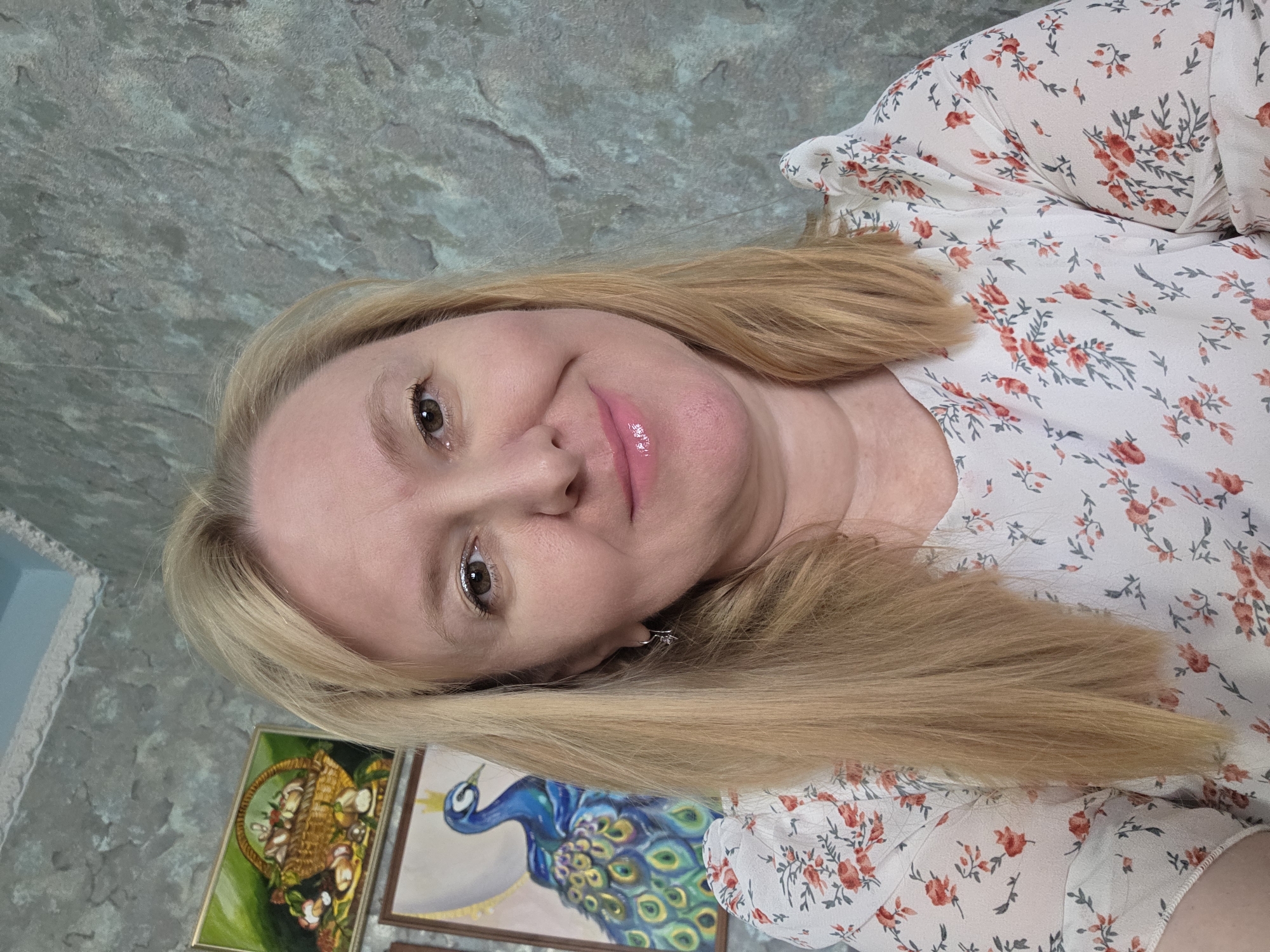 The image is a selfie taken indoors, likely in a residential setting in Novocheboksarsk, Russia. A blonde woman with fair skin and pink lipstick smiles at the camera. She is wearing a white blouse with a floral print of red flowers and green leaves. Behind her, on the wall, are two framed paintings. The painting on the left depicts a basket of fruits and vegetables, while the painting on the right features a blue peacock with a fan of colorful feathers. The wall itself has a textured, gray finish. The lighting suggests an indoor environment, possibly illuminated by natural light from a window or artificial room lighting.