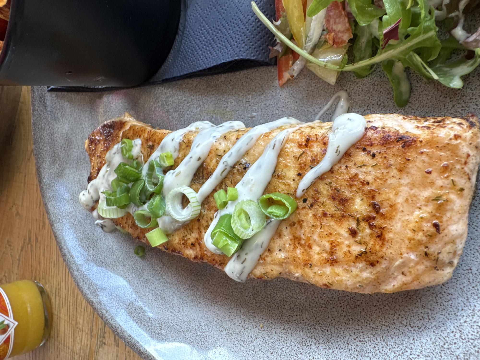 A pan-seared salmon fillet, approximately 20-25 cm in length, is centrally positioned on an oblong light grey speckled ceramic plate, angled slightly. The salmon exhibits a golden-brown crust with visible charring and small green specks, indicative of herbs such as dill or rosemary. It is drizzled lengthwise with a creamy white sauce, also containing small dark green flecks, suggesting a dill or herb-infused condiment. Generous portions of freshly chopped bright green scallions or spring onions are scattered over the top of the salmon. To the immediate right of the salmon, a small portion of mixed green salad is visible, containing arugula, red and yellow bell pepper strips, and possibly other leafy greens, lightly coated in a dressing. In the upper left background, a partial view of a small glass bottle containing a bright yellow liquid is visible on a light brown wooden tabletop. To the right background, a portion of a black cylindrical container, possibly a ramekin or small pot, rests on a dark grey or black textured napkin. The scene is set on a wooden surface, suggesting an indoor dining environment. No individuals are present in the image. The location is Unknown, Unknown.