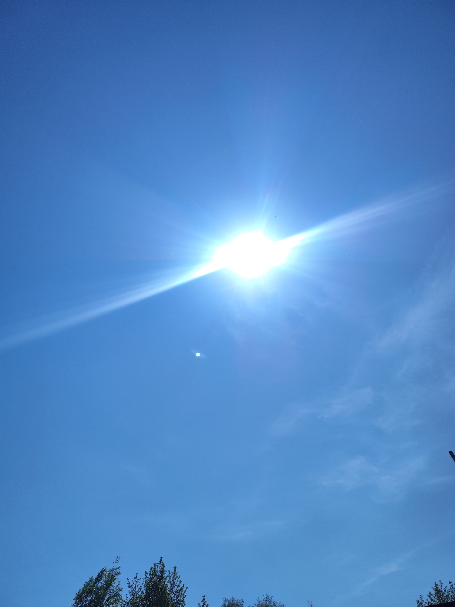 The image shows a bright sun in a clear, blue sky. There are some thin, wispy clouds in the lower right portion of the image. The tops of trees are visible at the bottom of the image, partially obscured by the low angle. There's a bright spot located beneath and to the left of the main sun, perhaps a reflection. The location is not possible to determine based on the provided information. No people or actions are discernible.