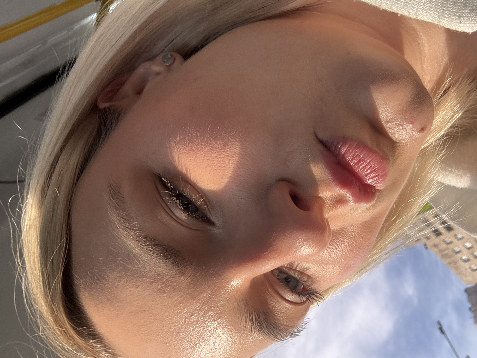A close-up, low-angle shot of a fair-skinned woman with blonde hair, positioned with her face tilted towards the camera. The subject has an earring on her left ear. The background shows a glimpse of a building and blue sky, suggesting an outdoor setting.