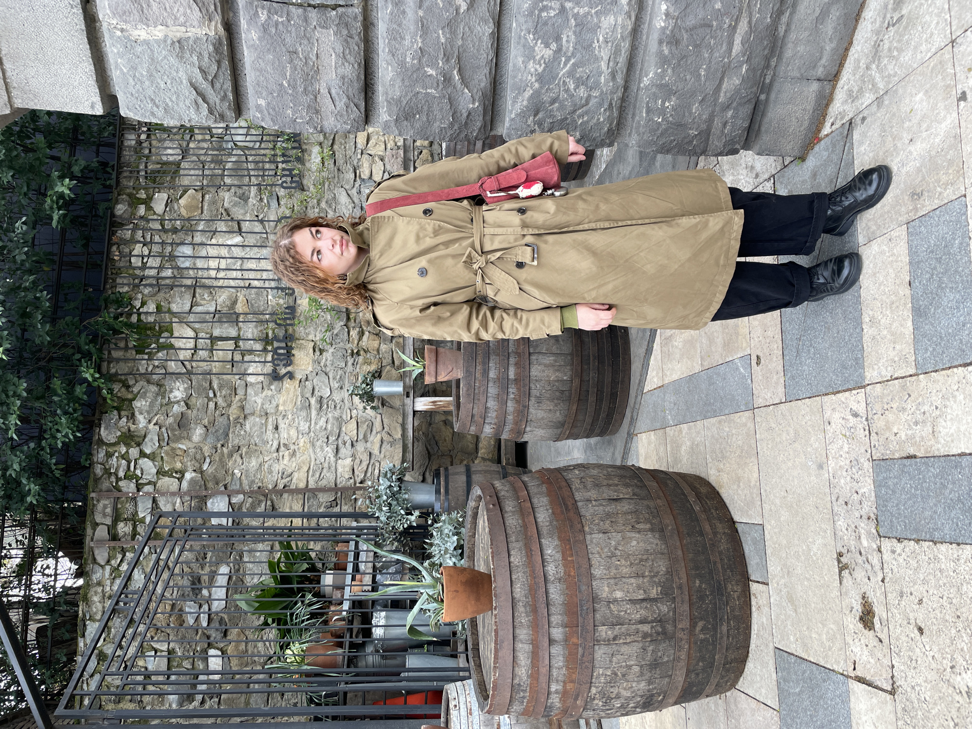 A medium shot captures a woman standing outdoors in Tbilisi, Georgia. She has curly hair and wears a tan trench coat, black pants, and black shoes. A red bag hangs over her shoulder. Behind her are two large wooden barrels, stone stairs, and a stone wall with a black metal fence. Potted plants are arranged near the barrels and fence. The ground is paved with light-colored tiles.