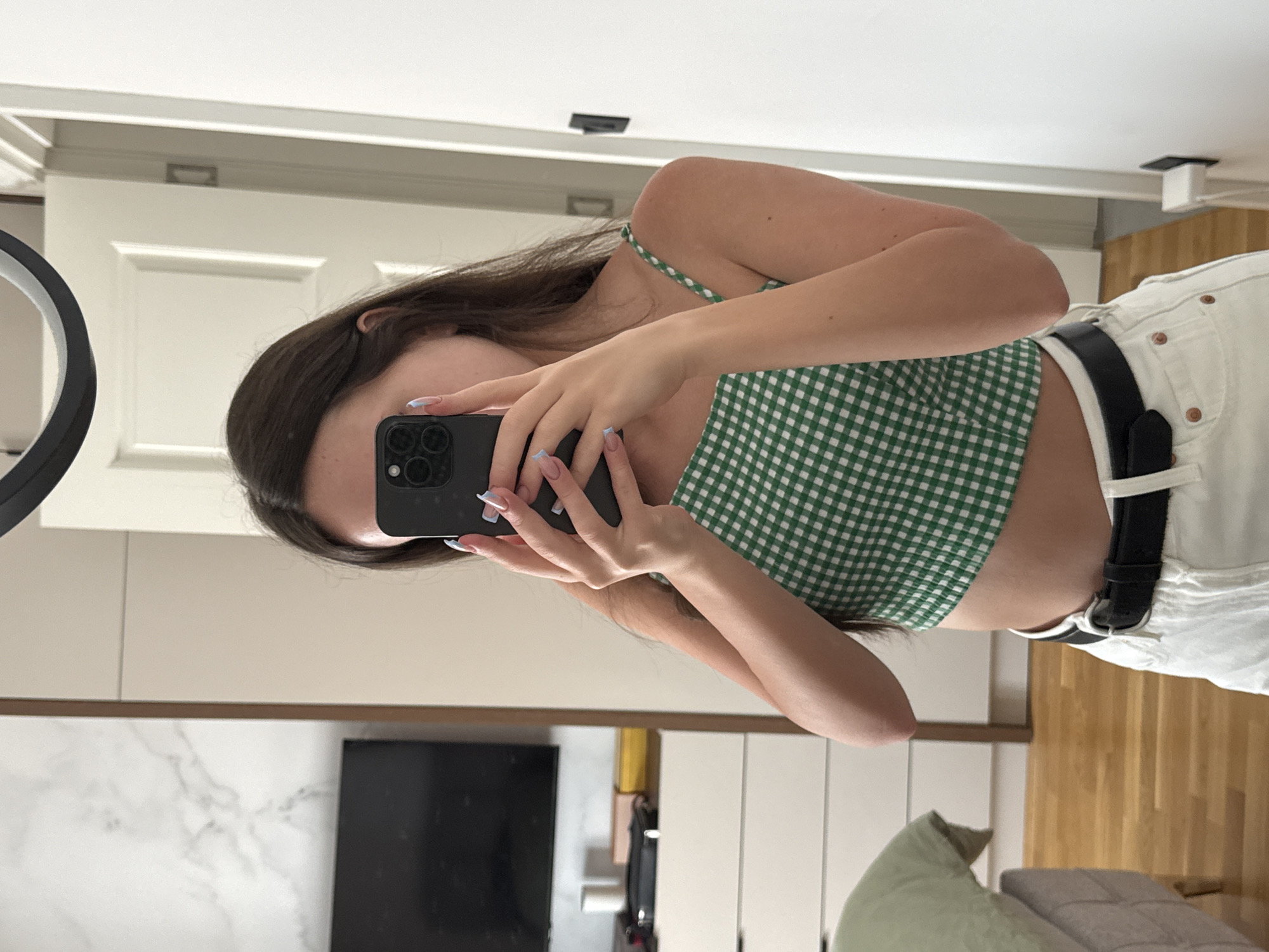 A person is taking a mirror selfie in what appears to be a bedroom. The person has long, dark hair and is wearing a green and white checkered crop top and white shorts with a black belt. They are holding a black smartphone in their right hand to take the picture. The background shows a white door, a white cabinet, and a wooden floor. There is also a glimpse of a television and some decorative pillows. The lighting suggests it is daytime.