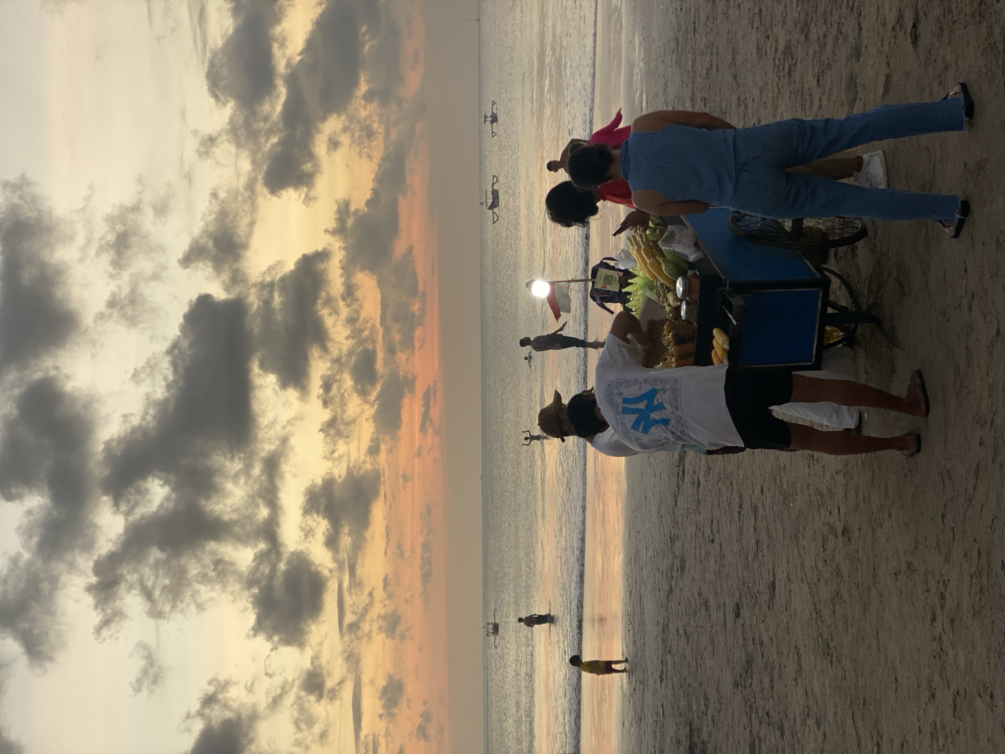 The image depicts a beach scene at sunset in Kuta Selatan, Indonesia. Several people are visible on the sandy shore and in the shallow water. In the foreground, a food cart is set up, laden with produce, likely corn. Two individuals are interacting with the cart, one wearing a blue t-shirt and jeans, and another in a white t-shirt with blue lettering and dark shorts. A third person, wearing a red garment, stands near the cart.
Further down the beach, other individuals are scattered. Two appear to be in the water, while others are walking along the shoreline. In the distance, two small boats or watercraft are visible on the calm sea.
The sky is dominated by dramatic clouds, tinged with the warm hues of sunset, indicating it is late afternoon or early evening. The overall mood of the scene is relaxed and typical of a beach setting during golden hour. The sand is coarse and appears wet near the water's edge.