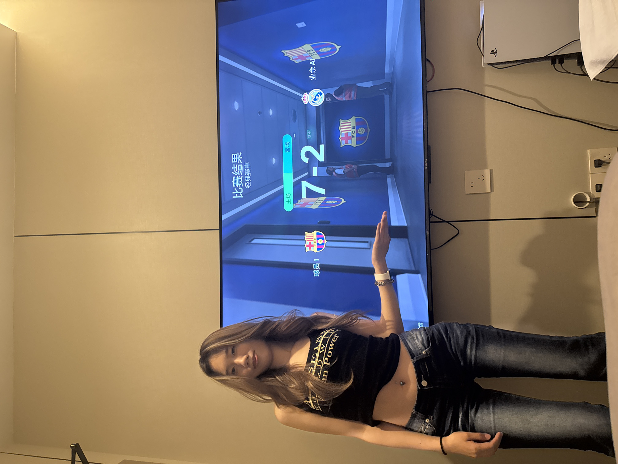 Here is a description of the image:
A young woman with long, wavy brown hair and wearing a black tank top with faded lettering and blue jeans stands in front of a large flat-screen television mounted on a light-colored wall. Her right arm is raised, palm facing forward. She is standing indoors.
On the television screen, a soccer game scoreboard shows a score of 7-2. The team logos of FCB (likely FC Barcelona) are displayed, and text in Chinese is visible above and below the scores. Above the television is a PlayStation 5 console. Power outlets and cables are also visible. The background wall is plain and painted a neutral tone.
The location in the city of Liangjiang, China, is not evident from the image.
