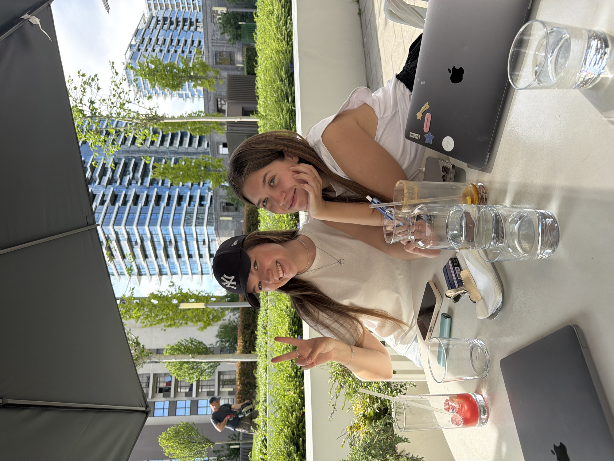 Two young women are seated at an outdoor table on a sunny day, likely in Belgrade, Serbia. The location is suggested by the modern architecture and landscaping in the background, characteristic of some areas of the city. The woman on the left wears a baseball cap and makes a peace sign with her fingers. The woman on the right is resting her head on her hand. Both are smiling and looking towards the camera. The table is covered with several empty glasses, a drink with red liquid, and two Apple laptops. A man pushing a cart is visible in the background. A large parasol partially covers the left side of the frame.