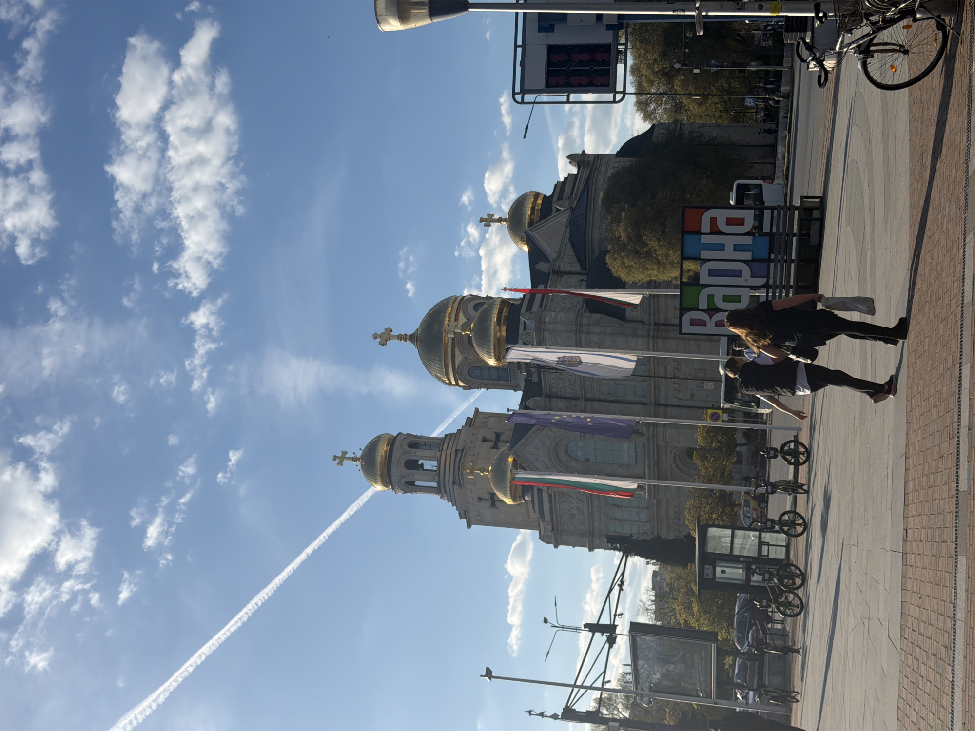 The image presents a vibrant urban scene in Varna, Bulgaria, dominated by the majestic Dormition of the Theotokos Cathedral, an Eastern Orthodox church, with its distinctive golden domes and crosses. Several flags, including the Bulgarian national flag (white, green, red horizontal stripes) and the European Union flag, are displayed on poles in front of the cathedral.
The sky is bright blue with scattered white clouds and a clear airplane contrail stretching across the upper left. The time of day appears to be mid-morning or afternoon, indicated by the strong sunlight and visible shadows.
In the foreground, a paved pedestrian area or street is visible, featuring a mix of light-colored and reddish-brown pavers. Two individuals are seen walking towards the left; one with long dark hair, dressed in dark attire, carries a white bag. Other urban elements include a street lamp post, a parked bicycle, and various street furniture and distant buildings. A prominent, colorful sign spells out "ВАРНА" (VARNA) in large Cyrillic letters, unequivocally identifying the location. The scene portrays a lively day in the city, centered around one of its most significant landmarks.