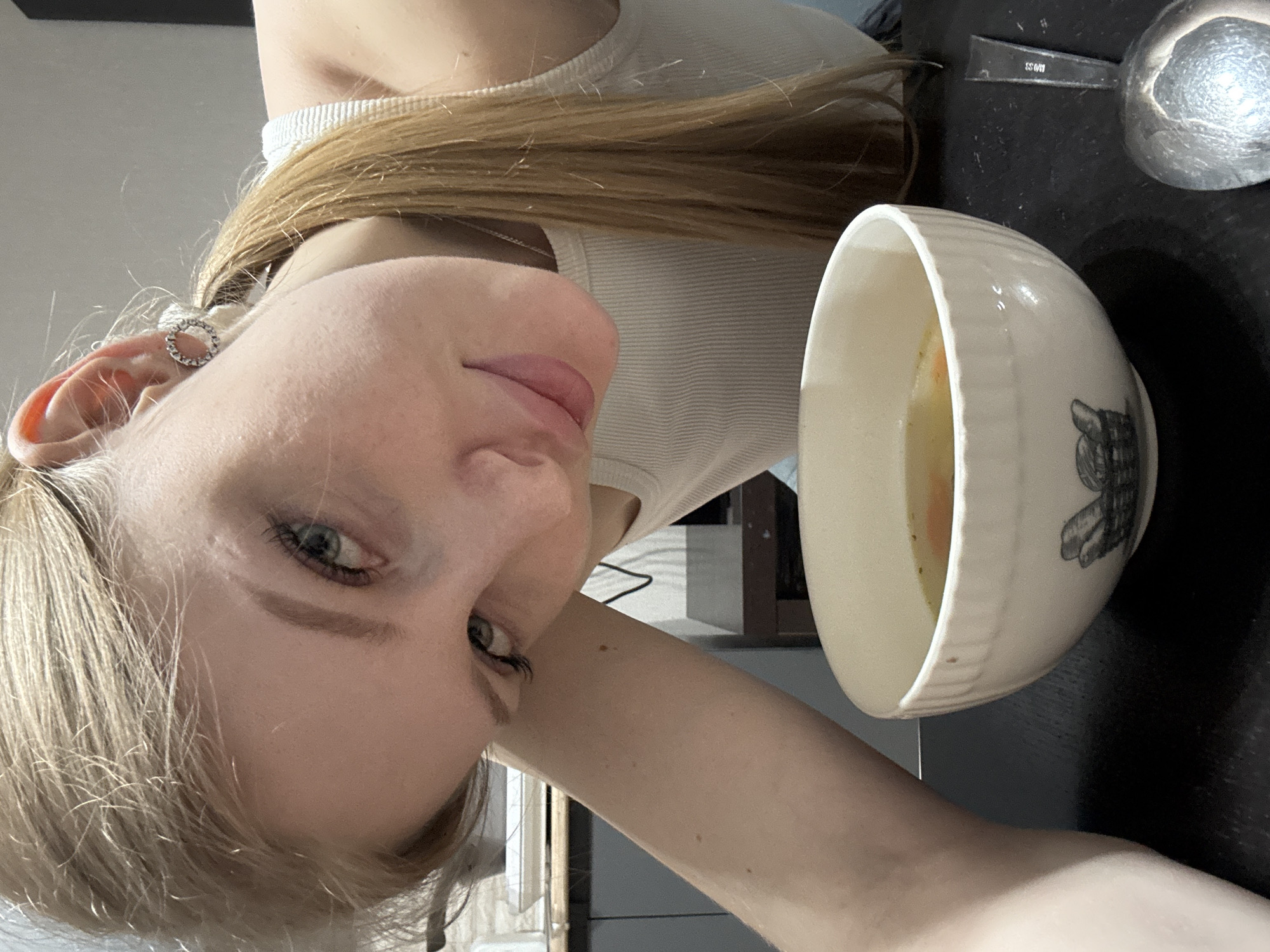 A young woman with blonde hair and blue eyes is holding a bowl of soup. She is wearing a white tank top and an earring in her left ear. A spoon is on the table next to the bowl. The image was taken indoors. I have no information regarding the location in the city of Perm, Russia.