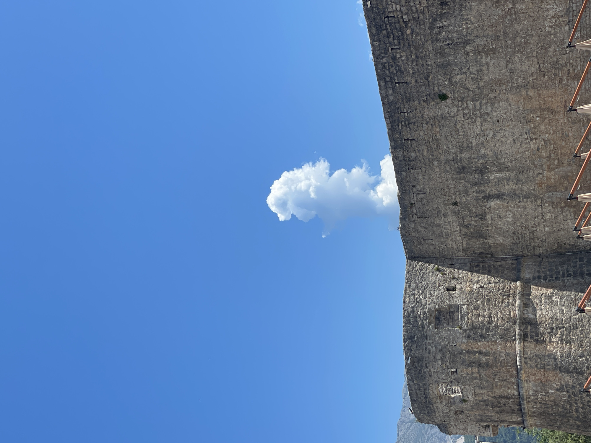 The image shows a bright blue sky with a single, large cumulus cloud in the center, shaped somewhat like a human figure. In the foreground, there is a portion of a stone fortification, likely a wall or tower, constructed from rough, grey stones. The fortification has several small openings and is casting a shadow. Below the fortification, the tops of several umbrella-like structures are visible, suggesting an outdoor seating area. In the background, distant mountains can be seen on the left side.