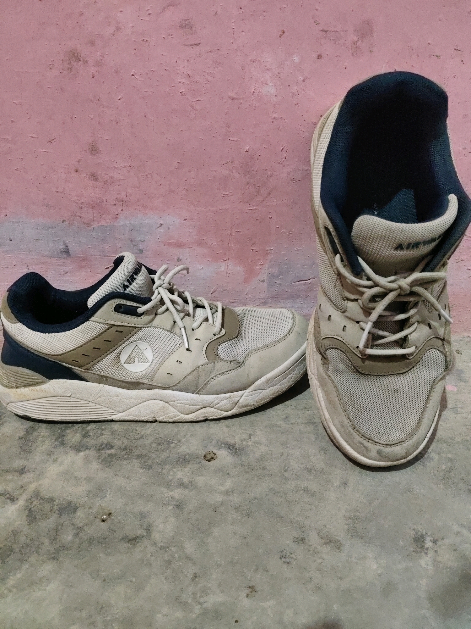The image shows a pair of used, light-colored athletic shoes placed on a concrete floor in front of a pinkish, textured wall. The left shoe is lying on its side, displaying its profile. The right shoe is standing upright, viewed from the front. Both shoes appear to be made of mesh and synthetic materials, with dark blue accents around the collar. The laces are visible and tied. The shoes show signs of wear and dirt. The environment suggests an indoor or covered outdoor space with a rustic aesthetic. There is no information in the image to indicate a location in Fajar Baru, Indonesia.