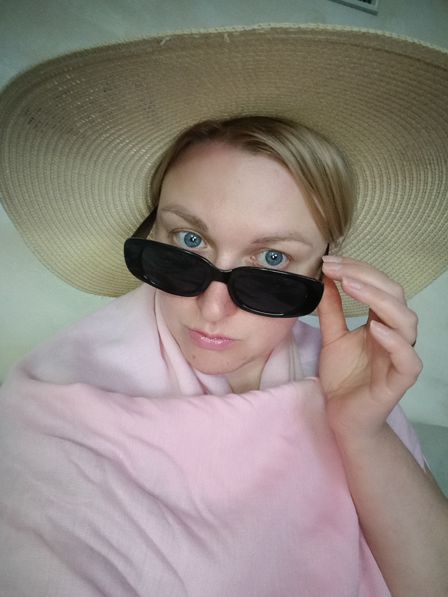 A fair-skinned woman with blue eyes is shown from a medium close-up perspective. She is wearing an oversized straw hat and black sunglasses, which she is adjusting with her right hand. A pink scarf is draped around her shoulders. The background appears to be a plain, light-colored wall. The location is Chaikovsky, Russia.