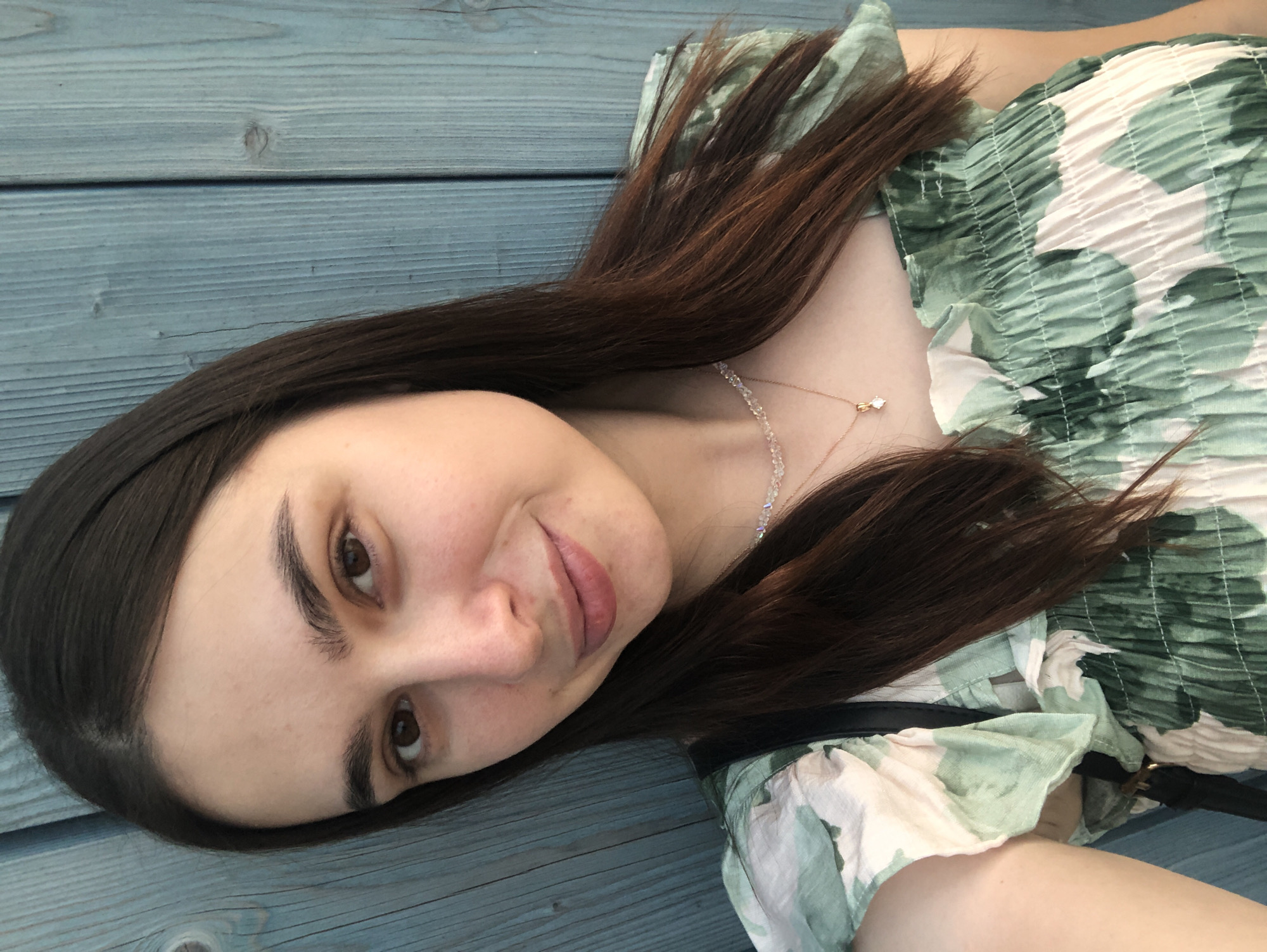 A young woman with long, dark brown hair is pictured from the chest up, against a backdrop of weathered blue wooden planks. She is looking directly at the camera with a slight smile. She is wearing a light-colored dress with a green floral pattern and a ruffled neckline. A delicate necklace with a small pendant is visible around her neck. The lighting suggests it is daytime, possibly outdoors, with the sun casting a soft light on her face. The scene appears to be a casual, everyday moment.