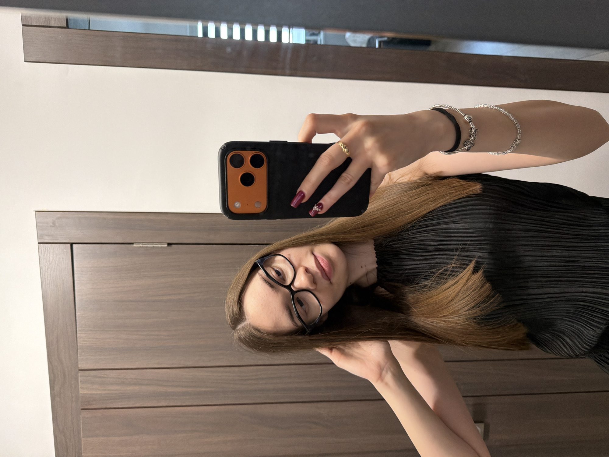 A young woman with long, straight brown hair and black-rimmed glasses takes a mirror selfie in an indoor setting, likely in Varna, Bulgaria. She is wearing a black pleated top and has a neutral to slightly smiling expression. Her right hand (left from the viewer's perspective) holds a black smartphone with a distinctive orange camera module, angling it towards the mirror. Her left hand (right from the viewer's perspective) is raised near her face, possibly adjusting her hair.
Notable accessories include a gold ring on her right ring finger, and her fingernails are painted with dark red (maroon) polish, featuring an embellishment on her right ring finger nail. On her left wrist, she wears two silver bracelets: one appears to be a delicate chain with a charm, and the other a tennis bracelet style with clear stones, alongside a black hair tie. The background reflected in the mirror shows a plain white wall and a section of dark wooden furniture or paneling. The mirror itself has a dark wooden frame visible at the top and bottom. There is no visible text in the image.