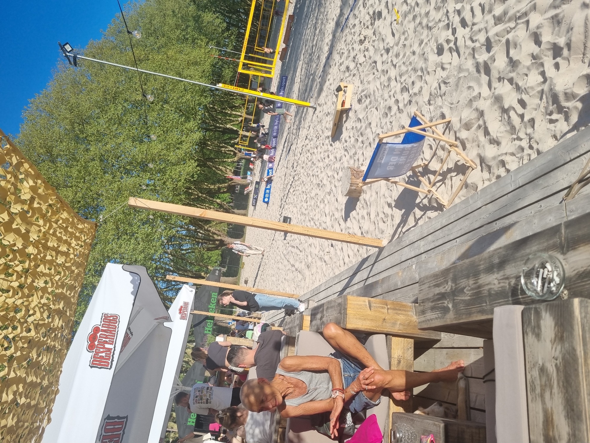 Here is a description of the image:
The photograph appears to be taken at an outdoor beach bar in Saarbrücken, Germany. The scene is brightly lit under what appears to be direct sunlight. In the foreground, there are individuals seated at wooden tables, one of whom is prominently displayed with their legs crossed. Other people are visible around the bar area, enjoying the ambiance. There are beverage umbrellas that say "Desperados" as well as "Heineken."
In the middle ground, a beach volleyball court is set up on the sand, with a volleyball net visible. In the background, a sparse crowd of people, possibly spectators or participants in the volleyball game, are spread along the sideline.
The environment suggests a relaxed beach vibe with light-colored sand covering the area surrounding the bar and volleyball court.