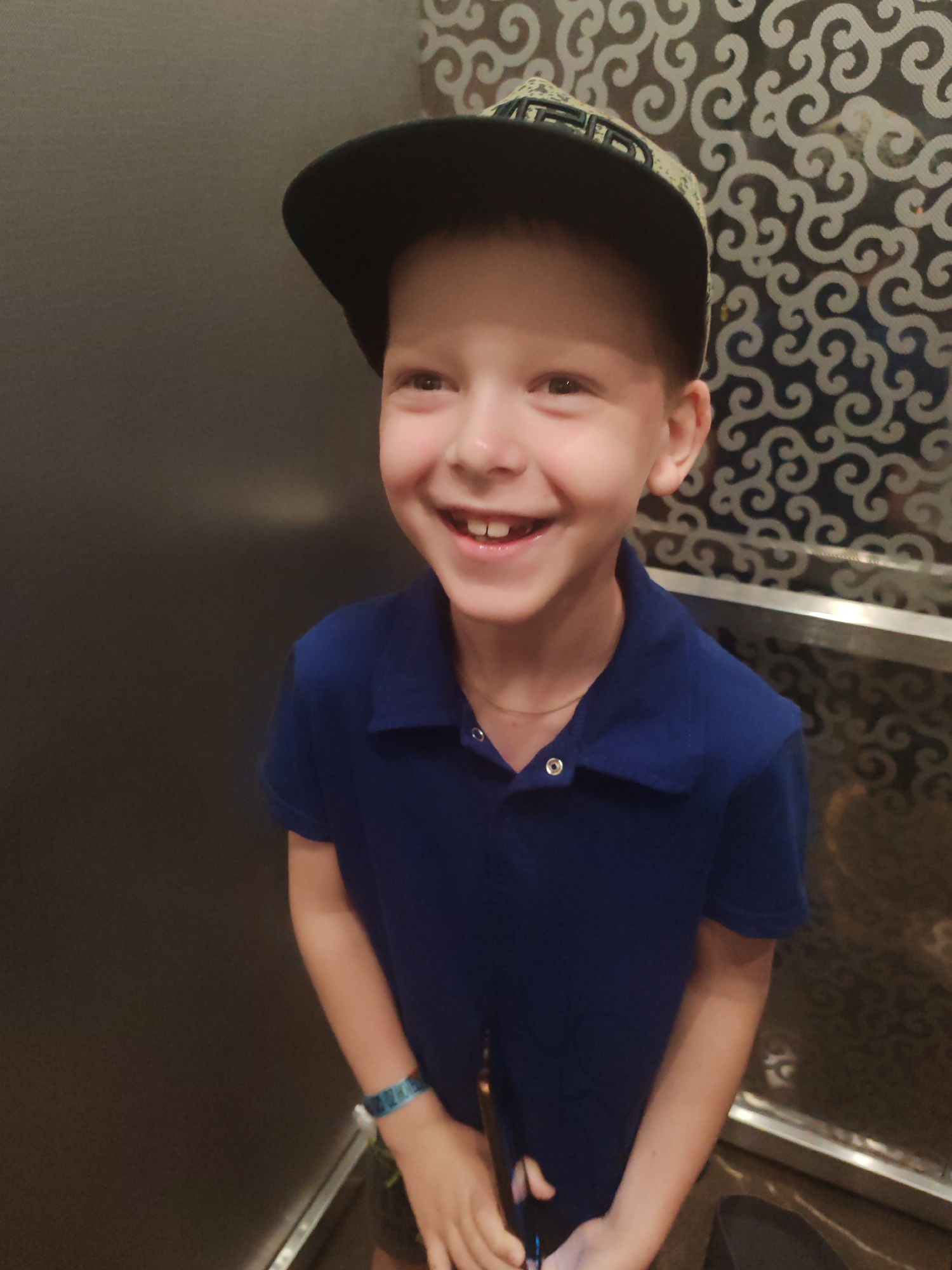 A young boy is smiling in an elevator. He is wearing a blue polo shirt and a baseball cap. He appears to be holding a phone in his hands. The elevator has a patterned wall. The location may be in the city of Alanya, Turkey.