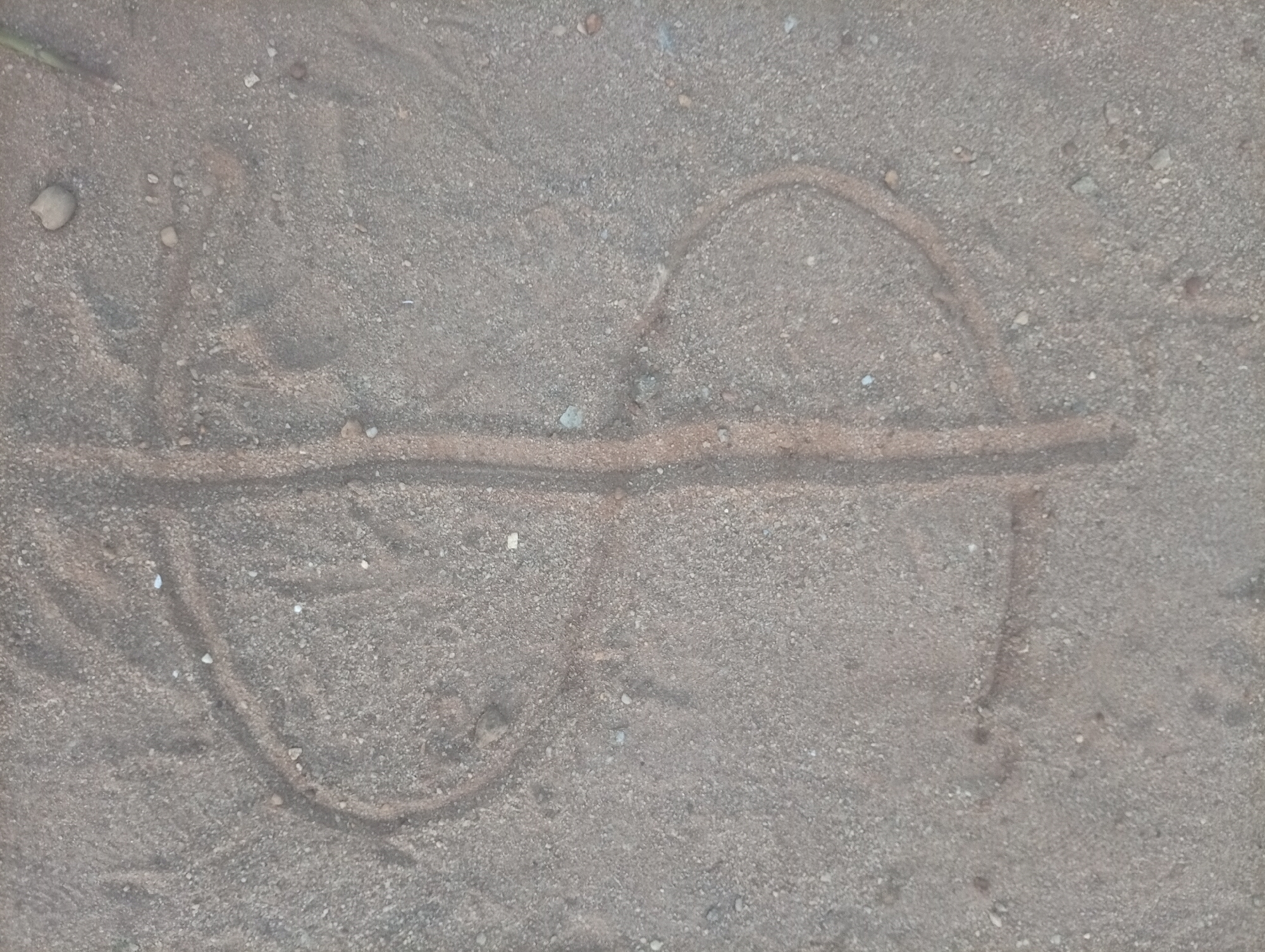 A drawing resembling the infinity symbol is etched in the sand in Ikorodu, Nigeria. The symbol is comprised of two incomplete circles connected by a horizontal line that extends beyond the right circle. The sand appears slightly damp. The location is sandy with small stones and pebbles scattered. No people or other objects are visible.