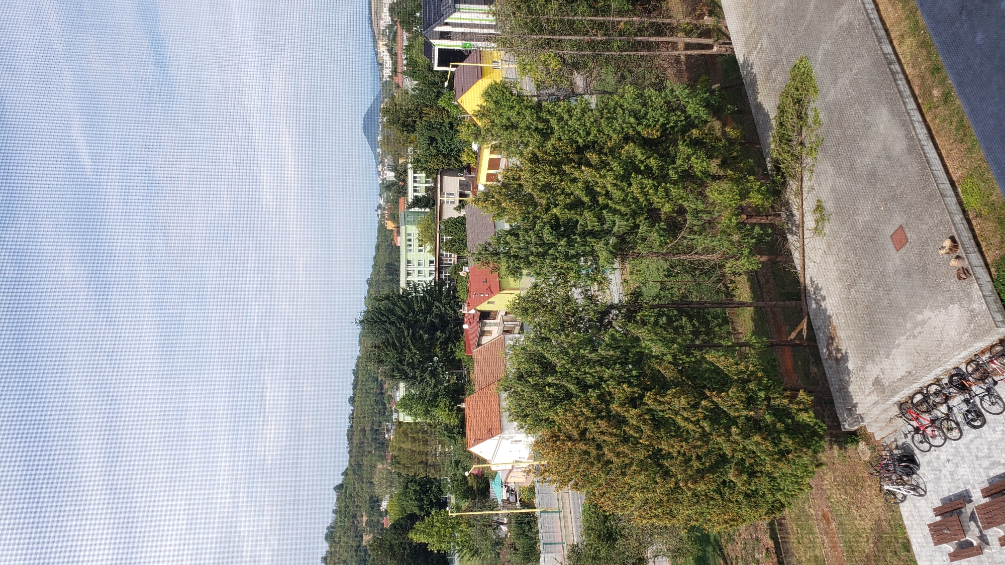 An elevated, slightly hazy view looks out over a suburban area in Prešov, Slovakia. In the foreground, a paved surface shows several bicycles parked near picnic tables. A fallen tree branch lies across the paved area. Behind this, a dense row of evergreen trees partially obscures a street and a cluster of houses with red and yellow tiled roofs. Further back, apartment buildings and more trees are visible against a backdrop of rolling hills under a cloudy sky. The image appears to have been taken through a window with a visible screen pattern.