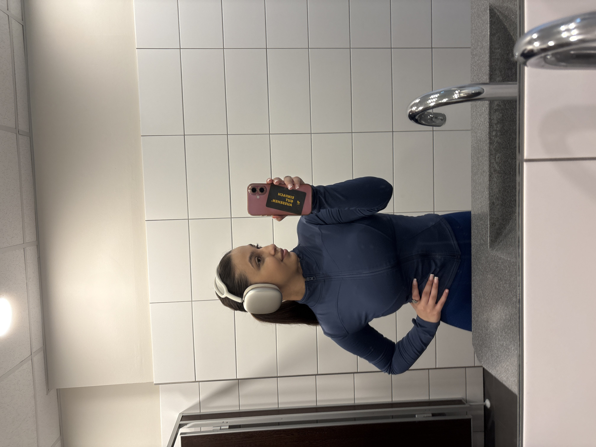 A woman wearing white headphones and a dark blue athletic top is taking a selfie in a restroom. She is holding a pink smartphone with a black card on the back. The restroom features white tiled walls and a sink with a chrome faucet. The location is likely within Mönchengladbach, Germany.