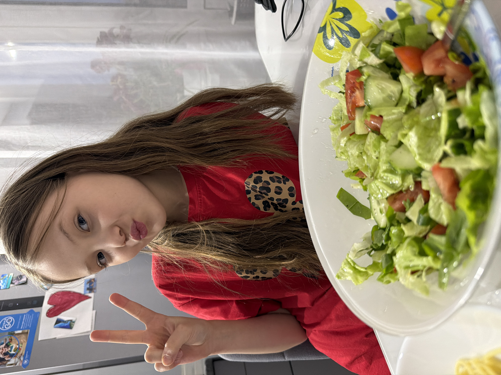 A fair-skinned young girl with long brown hair, wearing a red shirt with a leopard print, is sitting at a white table. She is making a peace sign with her right hand and pursing her lips. Beside her is a large bowl of salad containing lettuce, tomatoes, and cucumbers. The scene appears to be indoors, possibly in a kitchen or dining area. A window with sheer curtains is visible in the background. Magnets and papers are visible on a wall behind the girl. There is no information available in the image to confirm the location in the city of Chaikovsky, Russia.