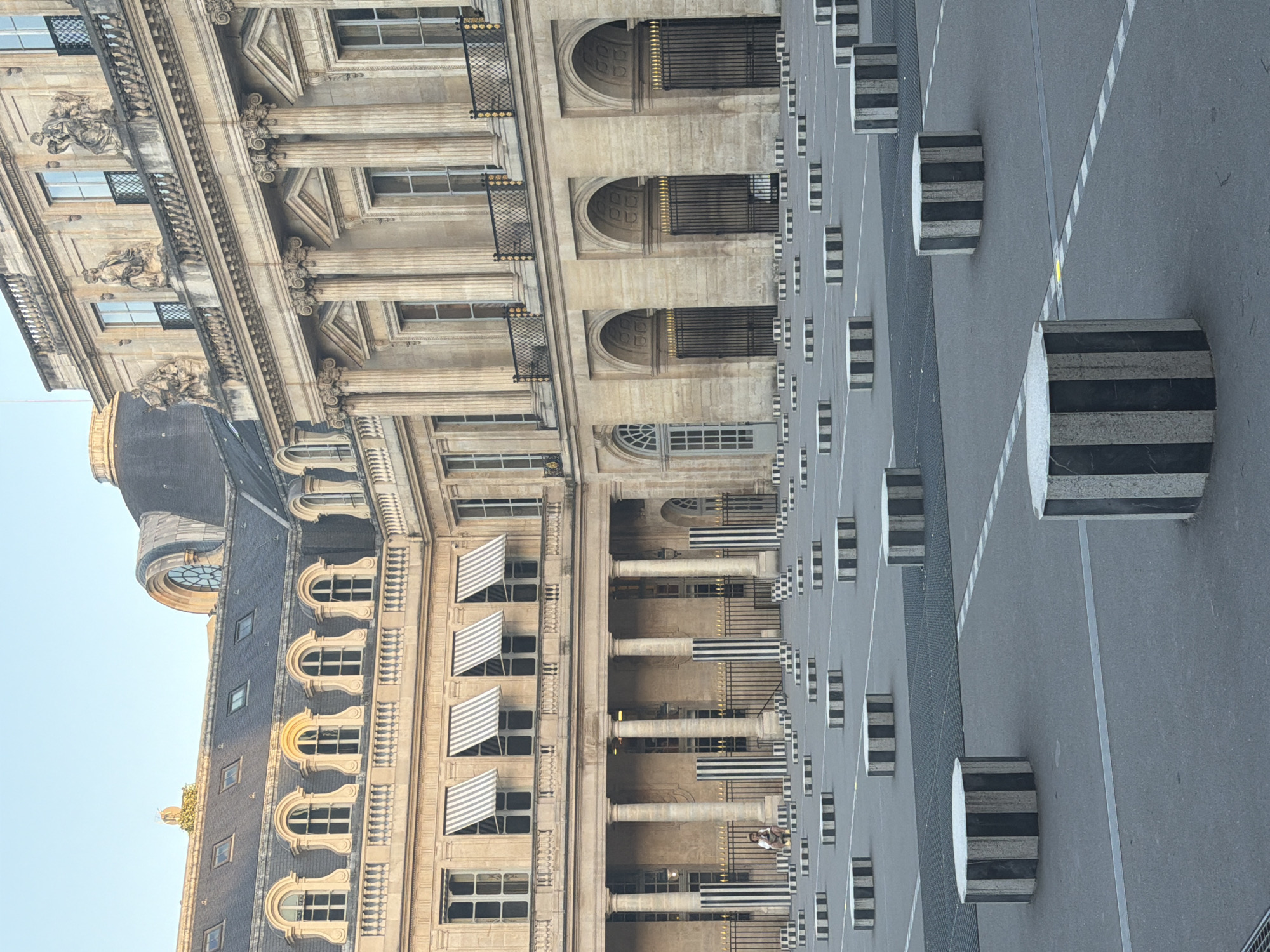 The image shows a street scene in Paris, France, with the Palais Royal visible on the left. The building is characterized by its ornate architecture, including columns, arches, and decorative elements. A portion of the roof and a clock tower are also visible. On the right side of the image, there is a street with striped bollards lining the edge of a walkway. The scene appears to be captured during the day under clear skies.