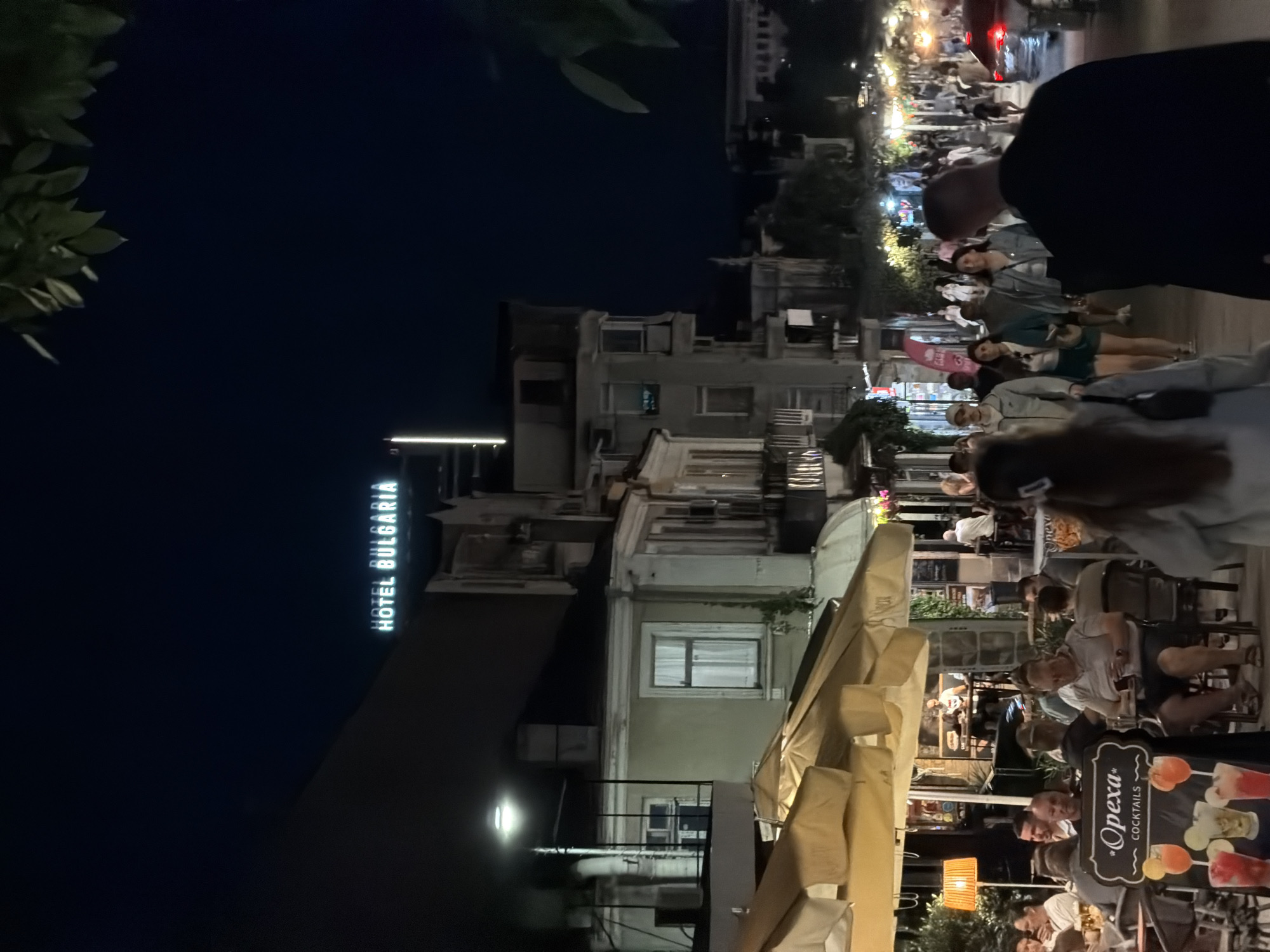 This is a nighttime, eye-level, medium shot of a bustling street scene in Burgas, Bulgaria. The "HOTEL BULGARIA" sign is visible illuminated against the dark sky. In the foreground, people are dining outdoors under awnings, with tables, chairs, and umbrellas. A sign for "*Opexa* COCKTAILS" with images of drinks is also present. Further down the street, a crowd of people walk along the pedestrianized area, with shops and buildings lining both sides. Streetlights and illuminated signs create a lively atmosphere. The street appears to be paved, and vegetation can be seen in the background. A car is visible in the distance on the right side of the frame. The overall impression is of a vibrant evening in a city center.